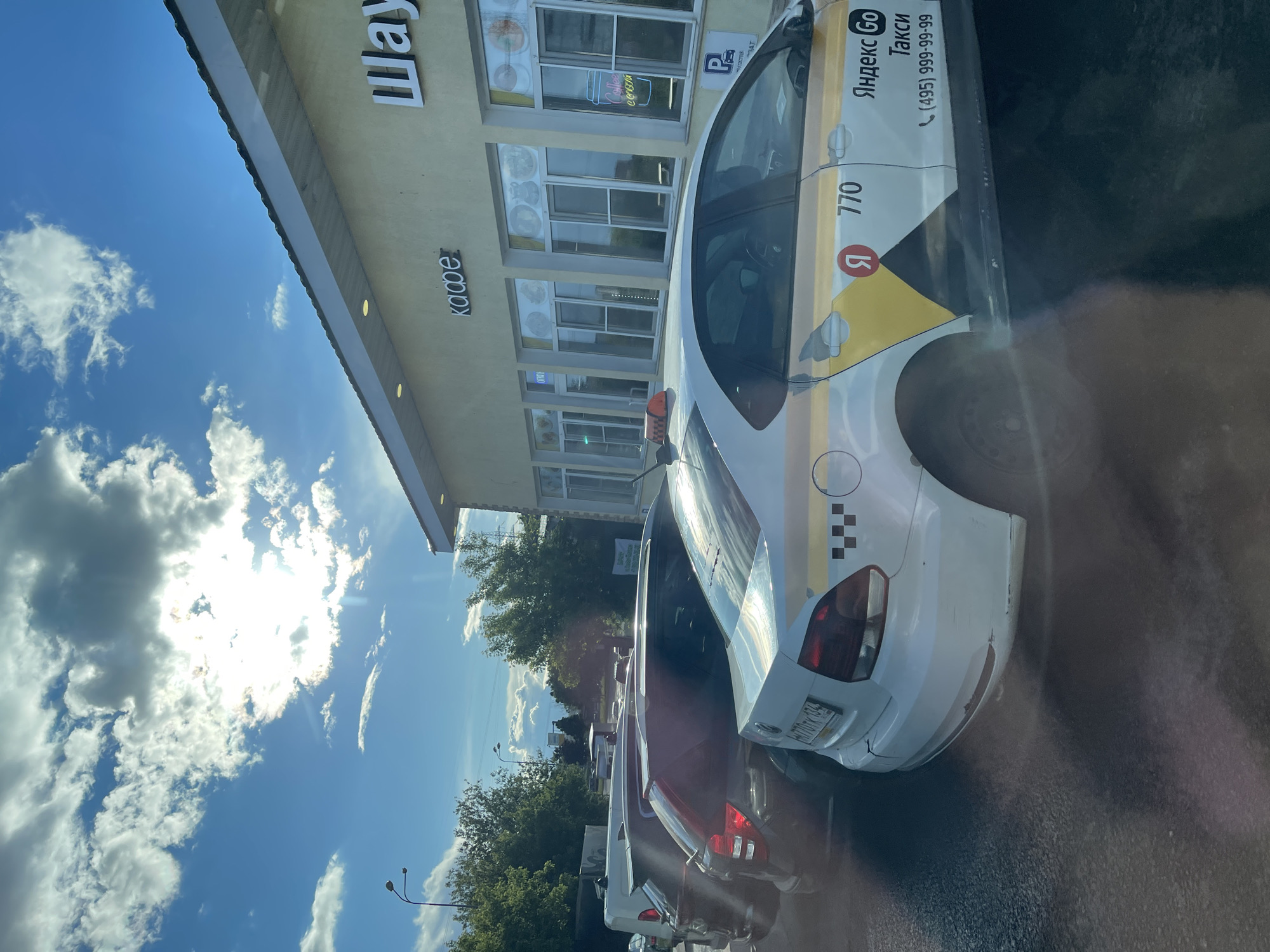 A daytime shot shows a street scene in Bittsa, Russia. Several cars, including a white Yandex taxi with the number 770 on its side and a taxi checker pattern, are parked along the street. A building with signs in Russian for "Shawarma" and "Coffee" is visible. The sky is blue with puffy white clouds. There is sunlight creating glare in the scene.
