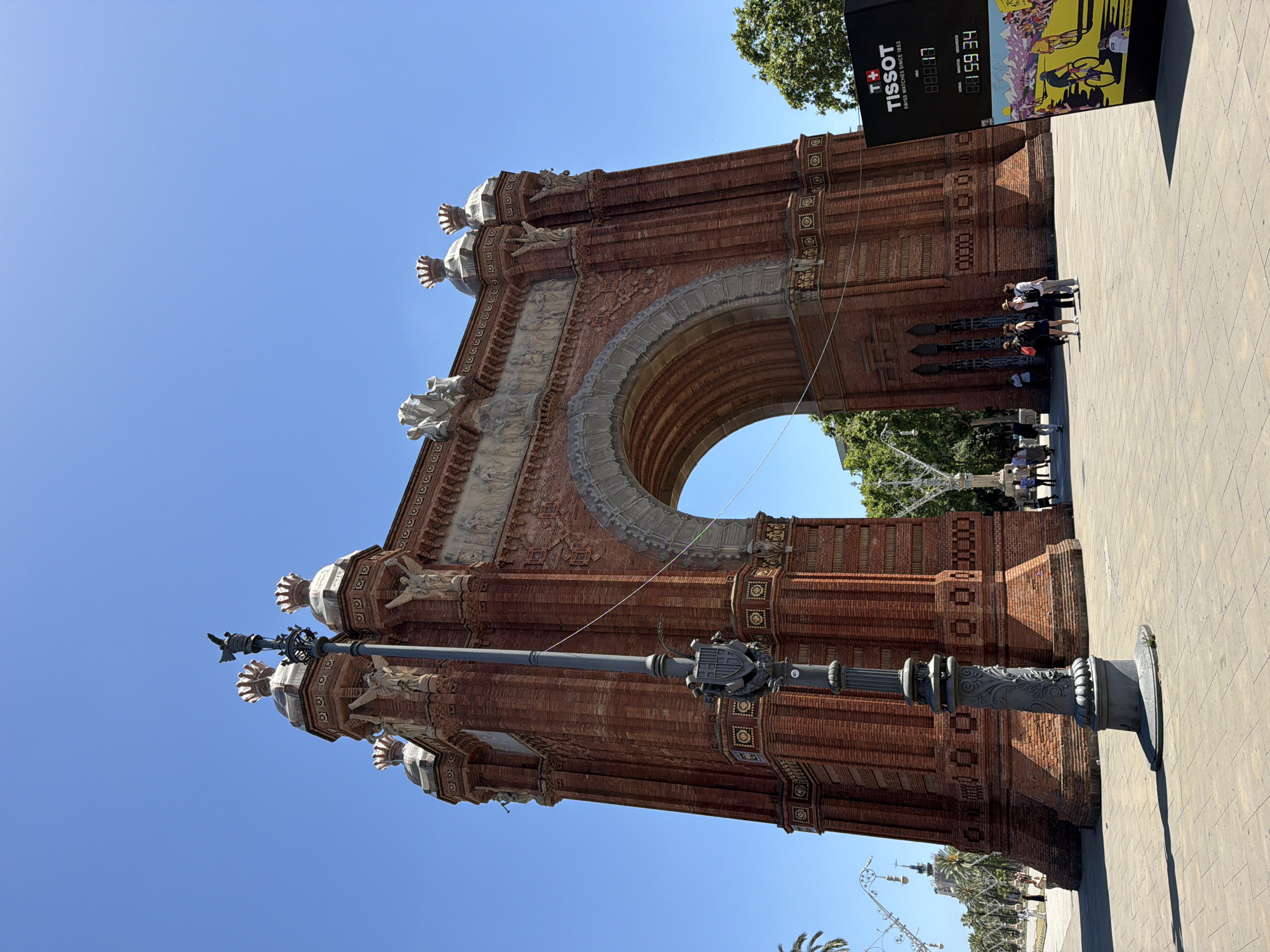 The image captures the Arc de Triomf in Barcelona, Spain, a prominent landmark situated at the entrance to the Parc de la Ciutadella. The monument, constructed from red brick with stone detailing, features a large central archway, ornate sculptures, and decorative elements at its apex. In the foreground, a lamppost with intricate carvings stands to the left of the arc. Several people are visible in the middle ground, walking and conversing in front of the monument. To the right, a digital display board with Tissot branding and a countdown timer is positioned. The background is dominated by a clear blue sky, with glimpses of trees and the city's architecture. The scene is bathed in bright sunlight, casting distinct shadows on the paved plaza.
