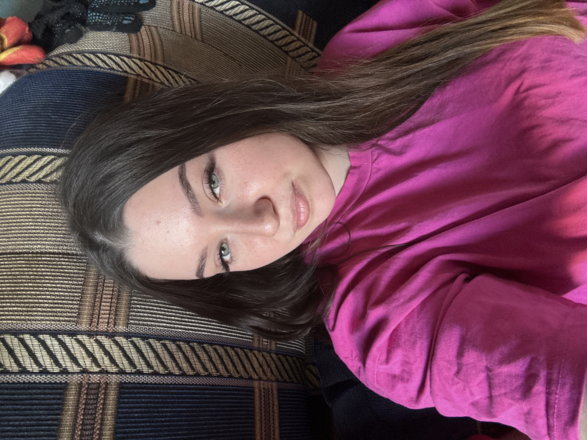 A medium shot shows a young woman with long, dark brown hair and green eyes. She is wearing a bright pink t-shirt and is looking towards the camera with a slight smile. The background appears to be a couch with a patterned fabric in shades of brown, beige, and dark blue. The image was captured in the city of Krasnokamsk, Russia, likely indoors.