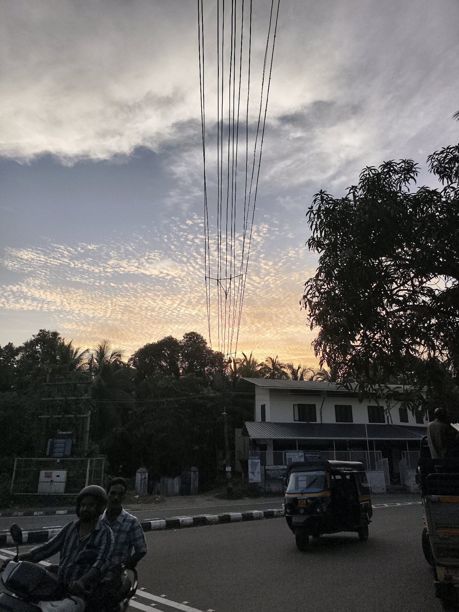 In the city of Mannoor Valavu, India, a street scene unfolds at dusk under a sky filled with scattered, luminous clouds. Utility wires stretch vertically across the frame, creating a stark contrast against the soft light. In the foreground, two men are riding on a motorcycle, the rider wearing a helmet. Behind them, the road curves, marked by a white and black striped median. Further back, an auto-rickshaw travels down the road. A modern, two-story building with a white facade and dark-framed windows sits to the right of the road, partially obscured by the dense foliage of a large tree. The background is a silhouette of trees, including palm trees, suggesting a tropical environment.