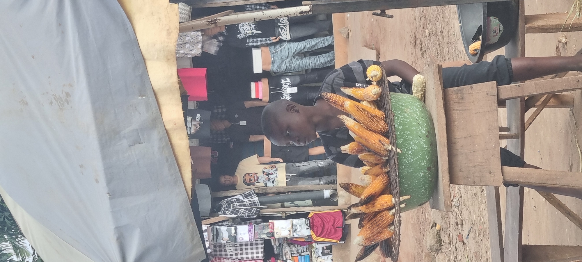 A young boy is roasting corn under a makeshift stall in Otta, Nigeria. The stall is covered with a faded grey tarp, and clothing racks displaying various shirts and pants are visible behind him. The boy is wearing a striped t-shirt and dark pants. In front of him, a metal rack holding several ears of roasted corn is placed over a large green bowl. Another ear of corn is visible on a wooden table to the right, along with a black bowl containing a partially roasted ear of corn. The ground is dirt and sand, with some scattered rocks.