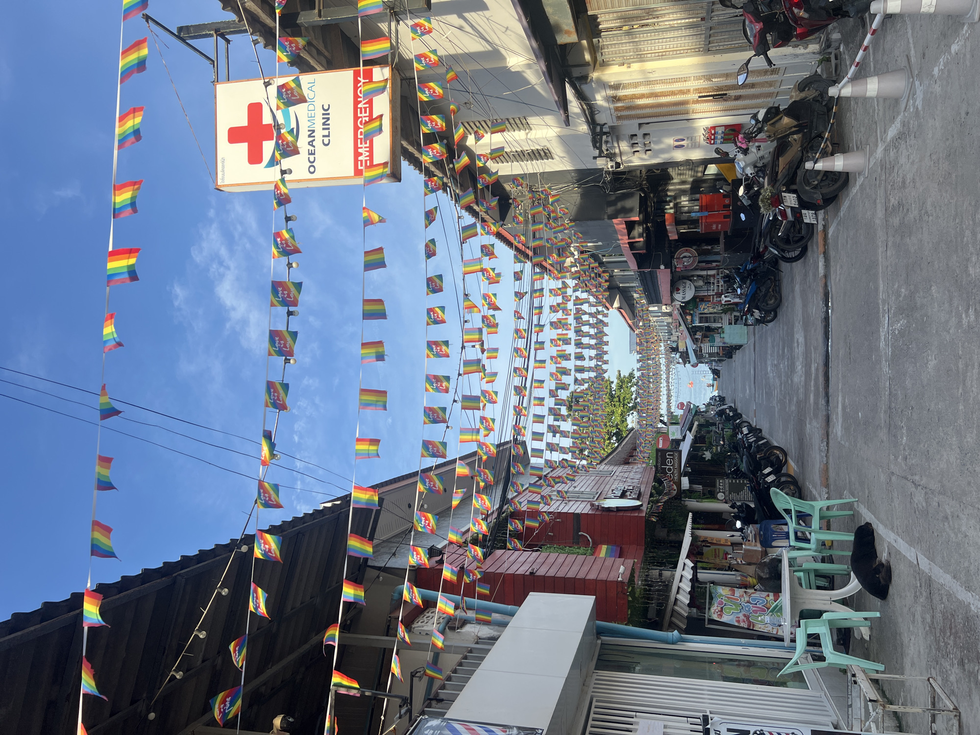 The image depicts a street scene in Ban Tai Subdistrict, Thailand, adorned with numerous rainbow-colored flags strung across the road, suggestive of a pride celebration or a LGBTQ+-friendly area. The street is lined with buildings on both sides, featuring shops and what appears to be a medical clinic. Several motorcycles are parked along the sides of the street. A black dog is sleeping on the sidewalk in front of a small table and chairs. The sky is blue with scattered clouds. The overall impression is of a commercial street in a tropical or subtropical location.