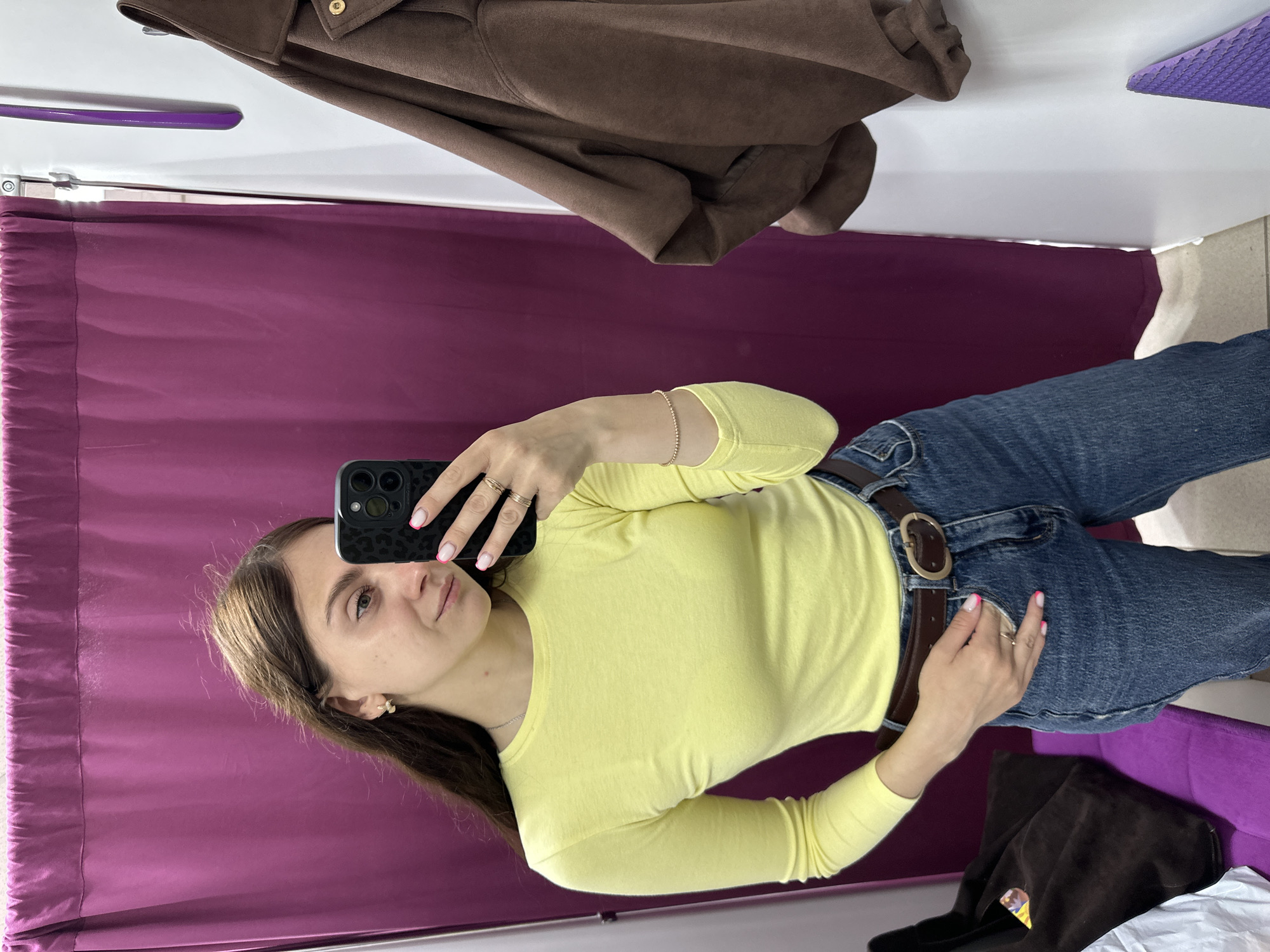 A woman takes a selfie in a dressing room. The woman is wearing a yellow long-sleeved shirt, blue jeans with a brown belt, and several rings. She has brown hair and fair skin. She is holding a black smartphone up to take the photo. In the background, there is a maroon curtain and a brown jacket hanging on a rack. The location of the photo is a dressing room, most likely in the city of Chaikovsky, Russia, although this cannot be determined definitively from the image alone.