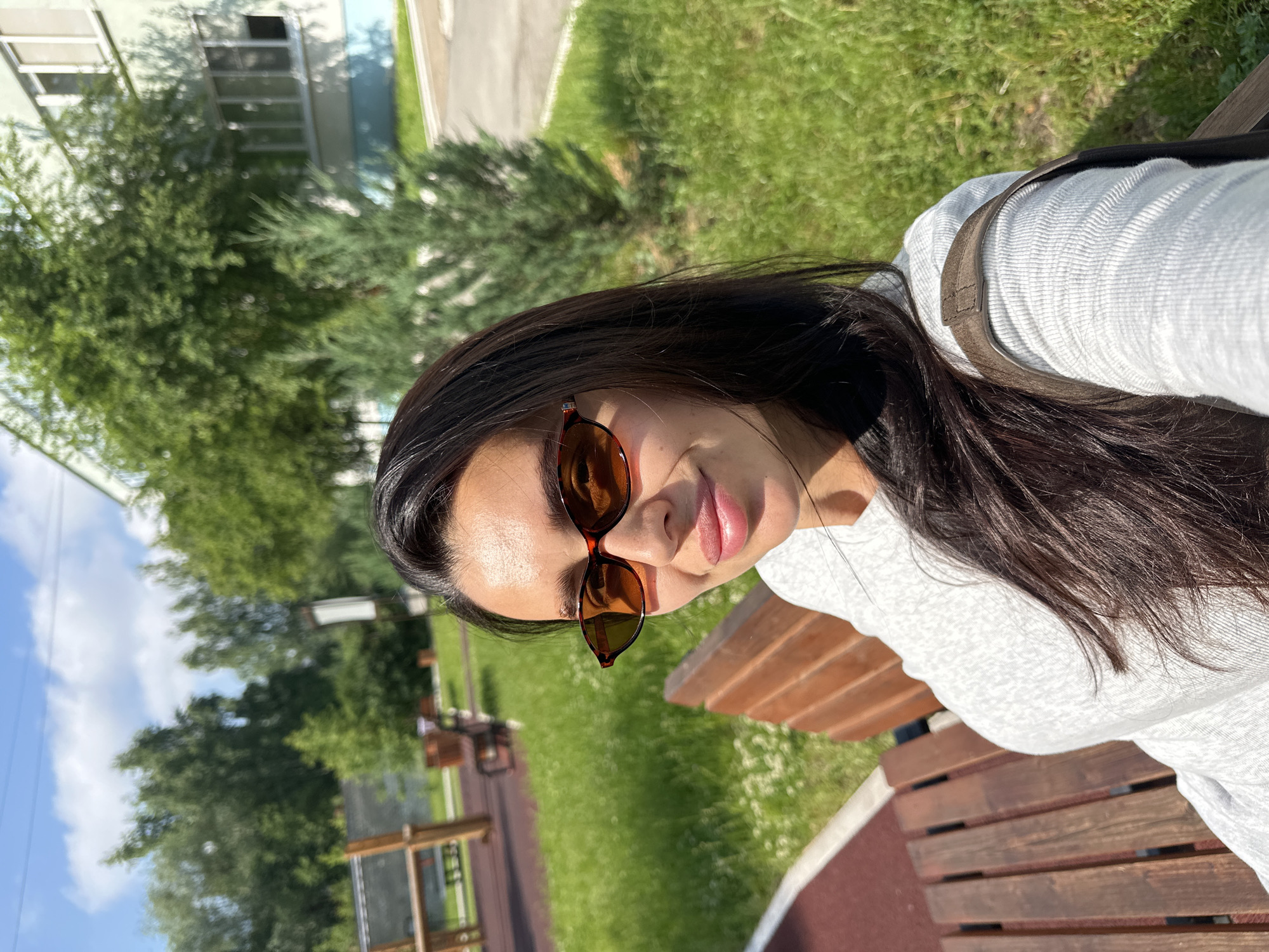 A young woman with long, dark hair and sunglasses is sitting on a wooden bench on a sunny day in Chaikovsky, Russia. She is wearing a light grey long-sleeved shirt and a brown strap is visible over her shoulder, suggesting she is carrying a bag. The background shows a grassy area, trees, and a building. To the left of the woman, a playground structure is partially visible. The overall scene depicts a relaxed outdoor setting.