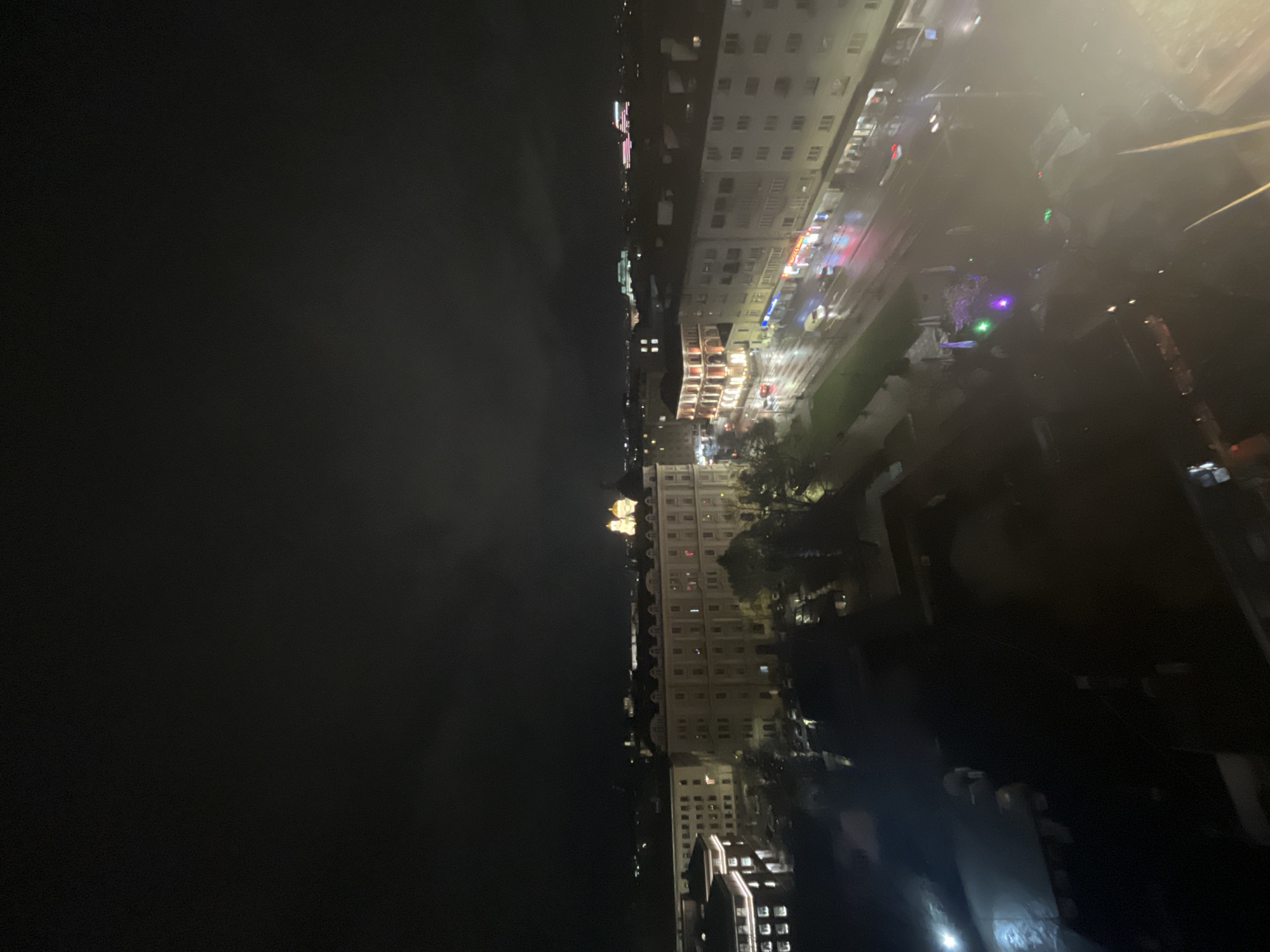 An aerial night view of Sofia, Bulgaria, reveals a cityscape illuminated by scattered lights. To the left of the frame, a dark expanse dominates, likely representing the night sky or a dark body of water. The city stretches from left to right across the middle of the frame, with buildings clustered together. The structures vary in height, and many show lit windows. Streets lined with light trails suggest vehicular movement. The light appears to be a mixture of warm and cool tones. Towards the top of the frame, there is a hazy or blurred effect, possibly from mist, fog, or atmospheric conditions. Trees appear scattered among the buildings.