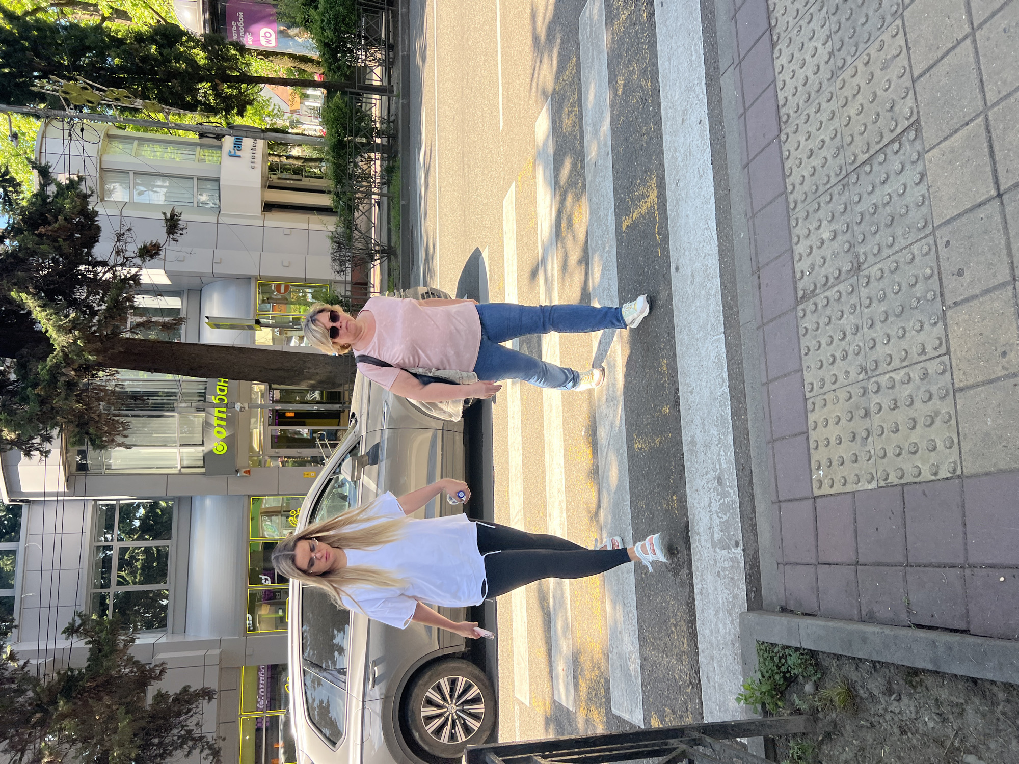 An outdoor, eye-level shot captures two women on a sunny day in Sochi, Russia, near a pedestrian crosswalk. In the foreground, a younger woman with long blonde hair, wearing a white t-shirt, black leggings, and white sneakers, stands on the edge of the zebra crossing, holding a clear bottle and a mobile phone. Behind her, slightly to the left, an older woman with blonde hair, wearing a pink t-shirt, blue jeans, and white sneakers, and sunglasses on her head, also stands on the crosswalk. Both women are looking towards the camera.
The setting is an urban street lined with trees and modern buildings. A light-colored car, possibly an SUV, is partially visible between the two women and the buildings. Shadows from trees and people are cast across the asphalt street and the white stripes of the crosswalk, indicating bright daylight, likely late morning or afternoon.
Notable visible text includes "Fami" and "Семья" (Family) on a white building sign, and "ОТП Банк" (OTP Bank) on a yellow-lit sign behind the younger woman. Further in the background, a purple billboard displays "Счастье это..." (Happiness is...) and the "WB" logo, likely for Wildberries. The sidewalk adjacent to the road features tactile paving. A dark metal railing is present in the lower foreground. The weather appears clear and sunny.
