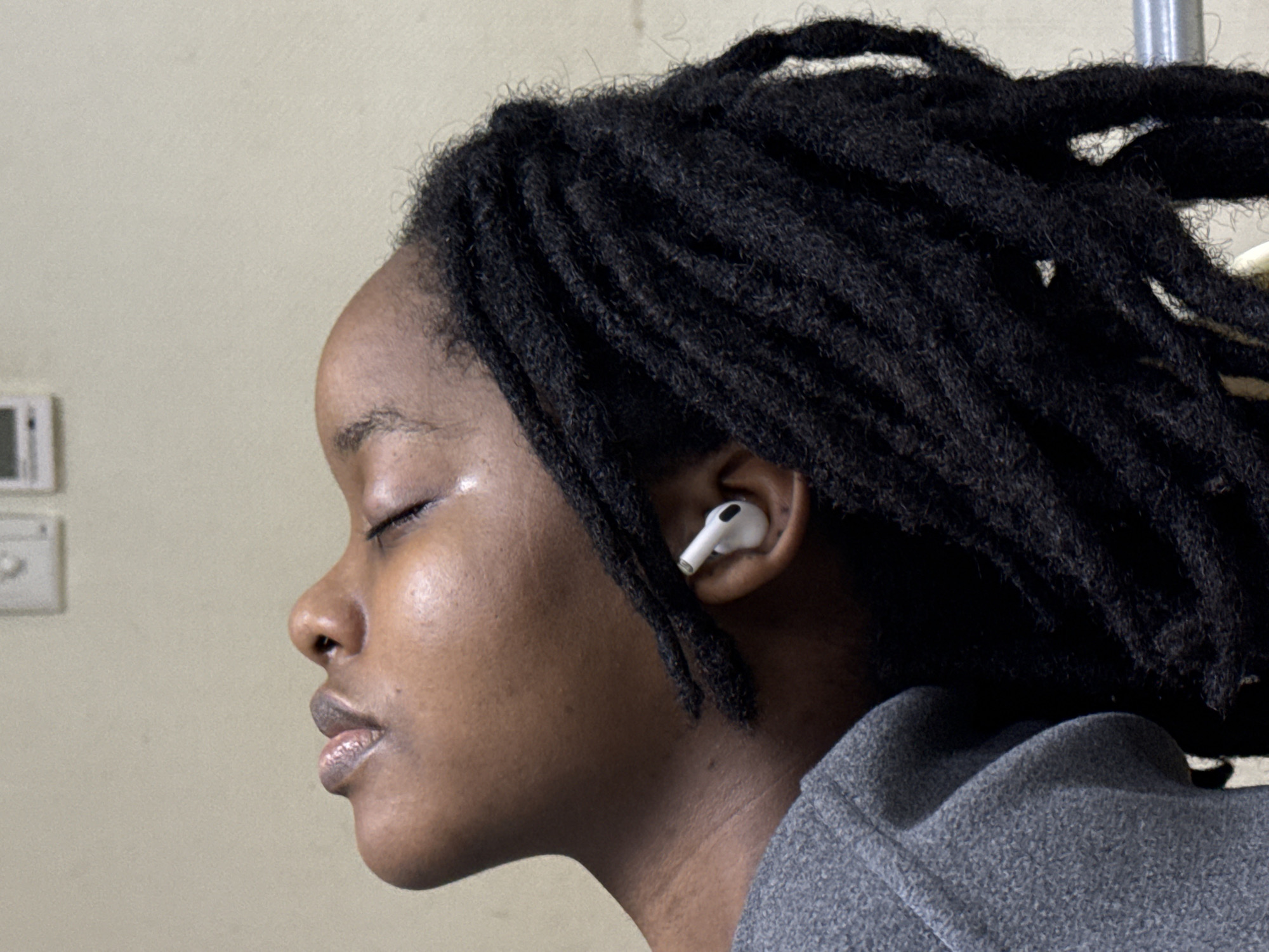 A medium shot captures a Black woman in profile, eyes closed, wearing an Apple AirPod in her ear. She has dreadlocks styled around her head. She is wearing a gray textured top. The background is a beige wall with a light switch and thermostat. The image lacks details to determine if it was taken in Yuzhong District, China.