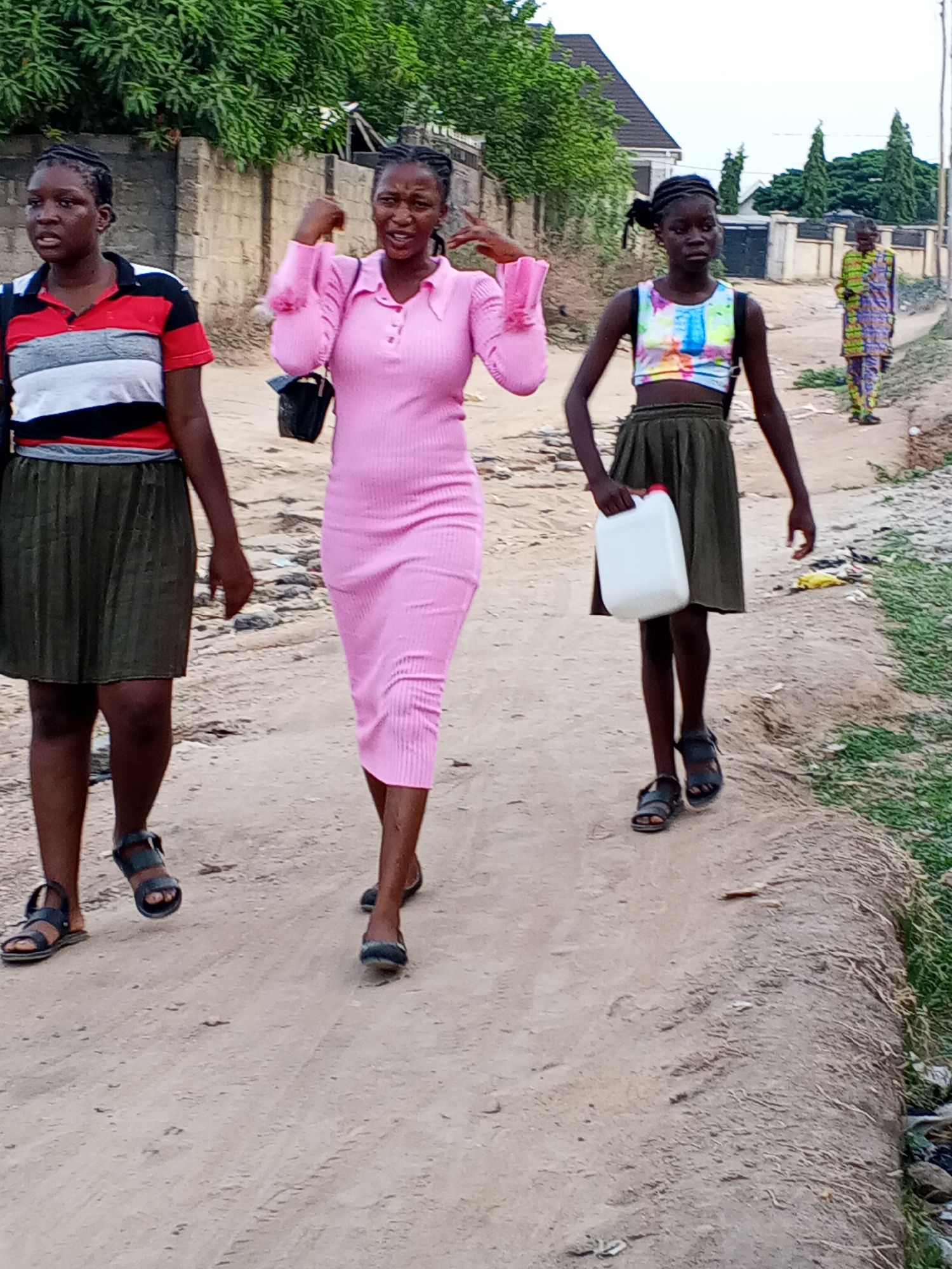 Three young women are walking on a dirt road in what appears to be a residential area of Unknown, Unknown. The woman on the left is wearing a striped shirt and a dark green skirt, and sandals. The woman in the middle is wearing a pink dress and flat shoes. She is gesturing with her hands. The woman on the right is wearing a colorful tank top and a dark green skirt, sandals, and is carrying a white plastic container. A fourth person in patterned clothing is further away on the road. The surroundings include buildings, greenery, and a dirt road.
