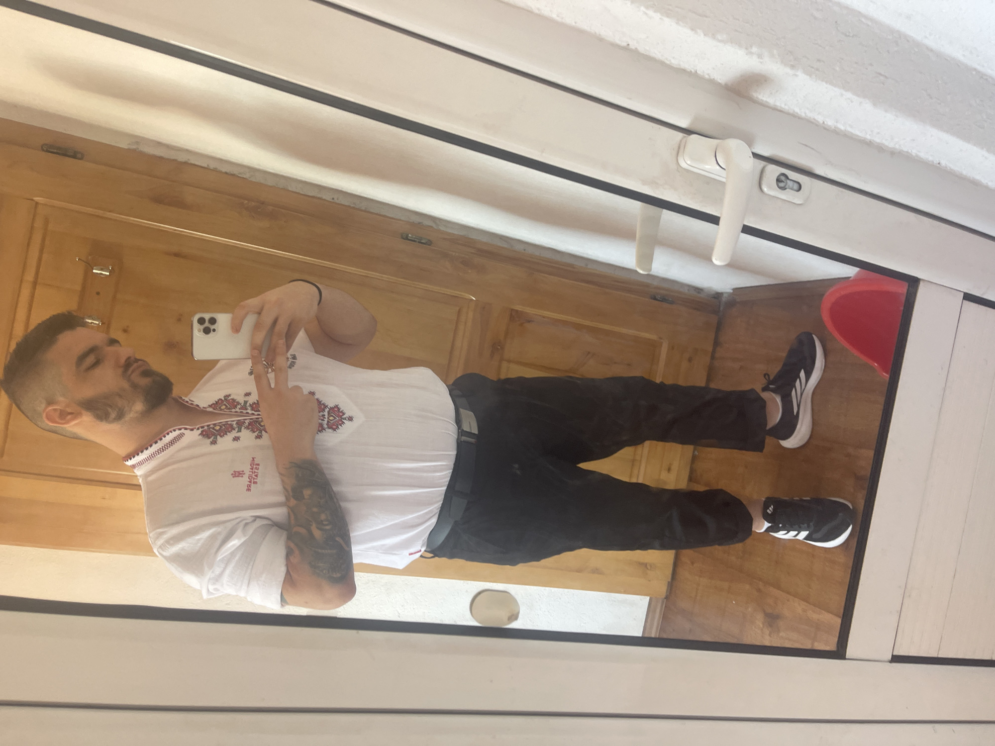 A man stands in front of a wooden door and a white door with a handle, taking a selfie in a mirror. He is wearing a white shirt with red embroidery on the collar and chest, black pants, and black and white sneakers. He has tattoos on his left arm. The background shows a wooden door and a white door frame with a handle. The floor is made of wood.
