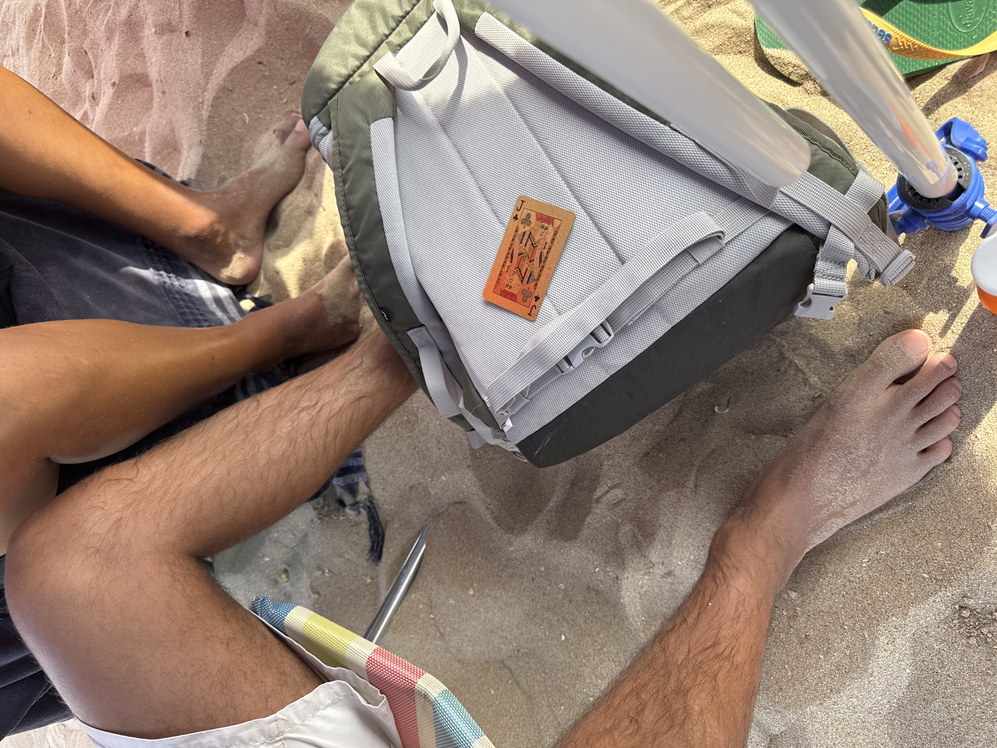 The image shows a close-up view of people's legs and feet on a sandy beach, likely in Costa da Caparica, Portugal, based on the context provided. The sand is light brown and appears somewhat damp in places, with some small shell fragments visible. In the foreground, a person's tanned leg with visible dark hair is seen resting on a striped beach chair, with white shorts visible. To the right, another person's legs and feet, covered in sand, are visible. A backpack, predominantly dark green and grey, is positioned in the center of the frame. A single playing card, featuring an illustration of a man (likely a Jack of Diamonds), is placed on the backpack. The background suggests other people are present, with more legs and sand visible. The lighting indicates it is daytime, with the sun casting shadows and highlighting textures in the sand and on the skin.