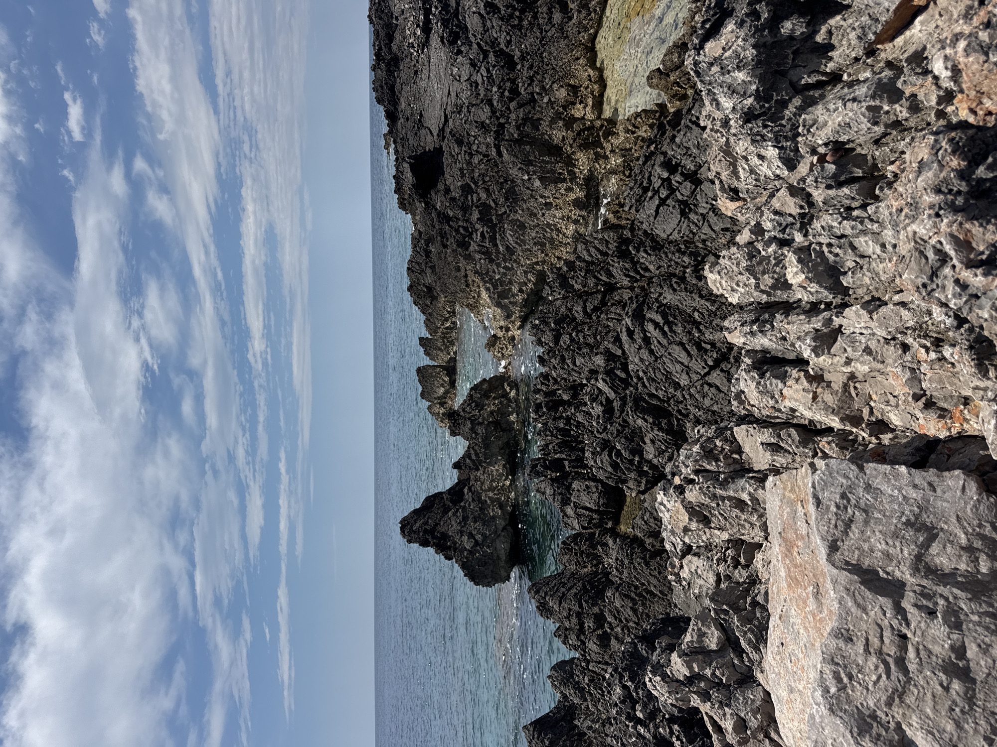 The image depicts a rocky coastline in Dubraljevina, Montenegro. The right side of the frame features a dark, jagged rock formation. To the left of the rocks is the Adriatic Sea, appearing calm and turquoise. The sky is visible on the left, showing a mix of blue and elongated white clouds. There are no people visible in the scene. The focus is on the natural landscape.