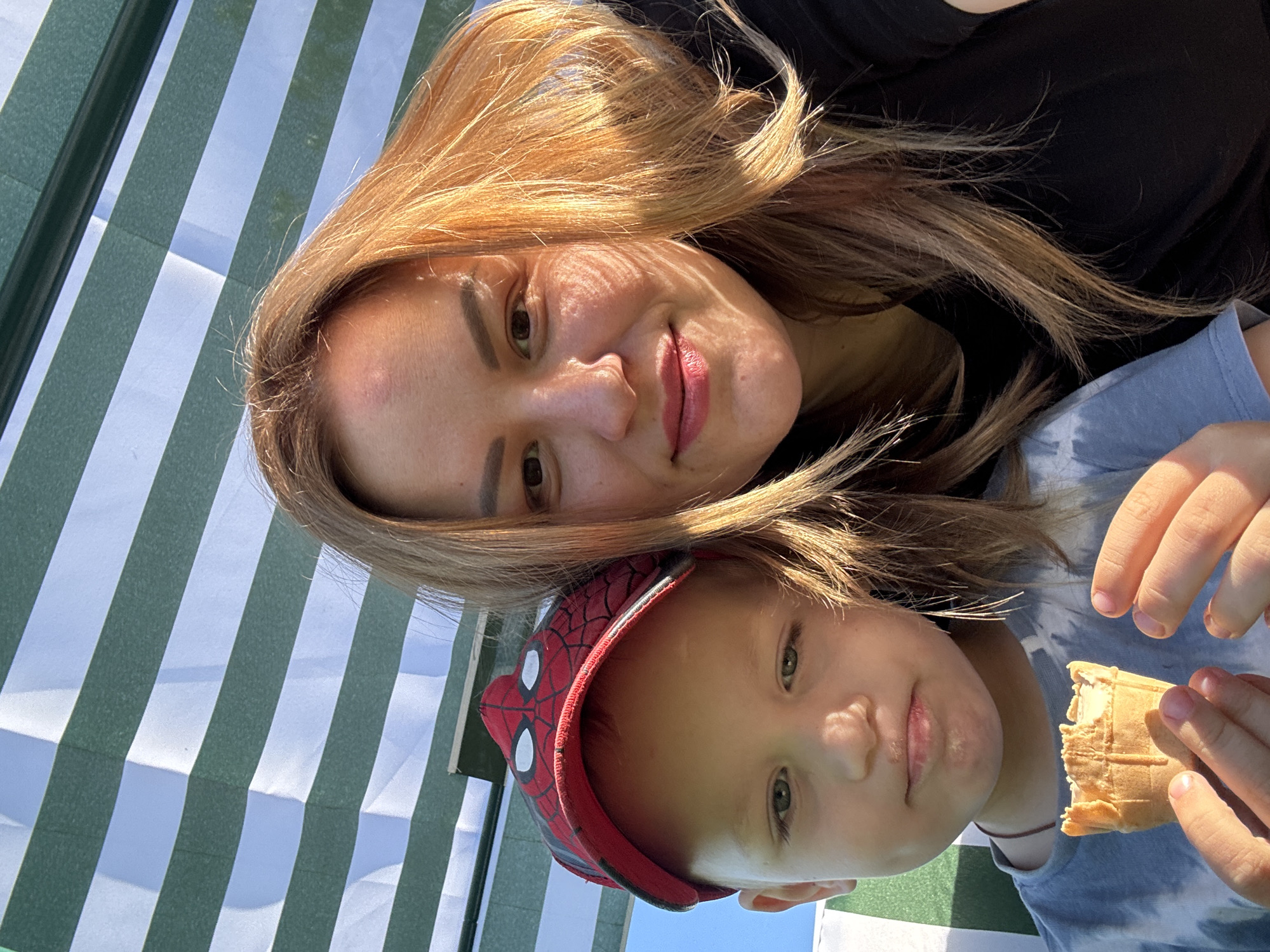 A medium-shot selfie shows a woman and a young boy outdoors, seated under a green and white striped awning. The woman is on the right, her face turned towards the camera, with golden-brown hair and light makeup. She is wearing a black t-shirt. The boy is on the left, wearing a red Spider-Man baseball cap and a blue tie-dye t-shirt. He is holding an ice cream cone with a bite taken out of it in his hands, looking downwards with a slightly wistful expression. Sunlight casts shadows on the woman's face. The background consists of the striped awning and a clear blue sky. The location is not identifiable as Chaikovsky, Russia, or any other specific city.