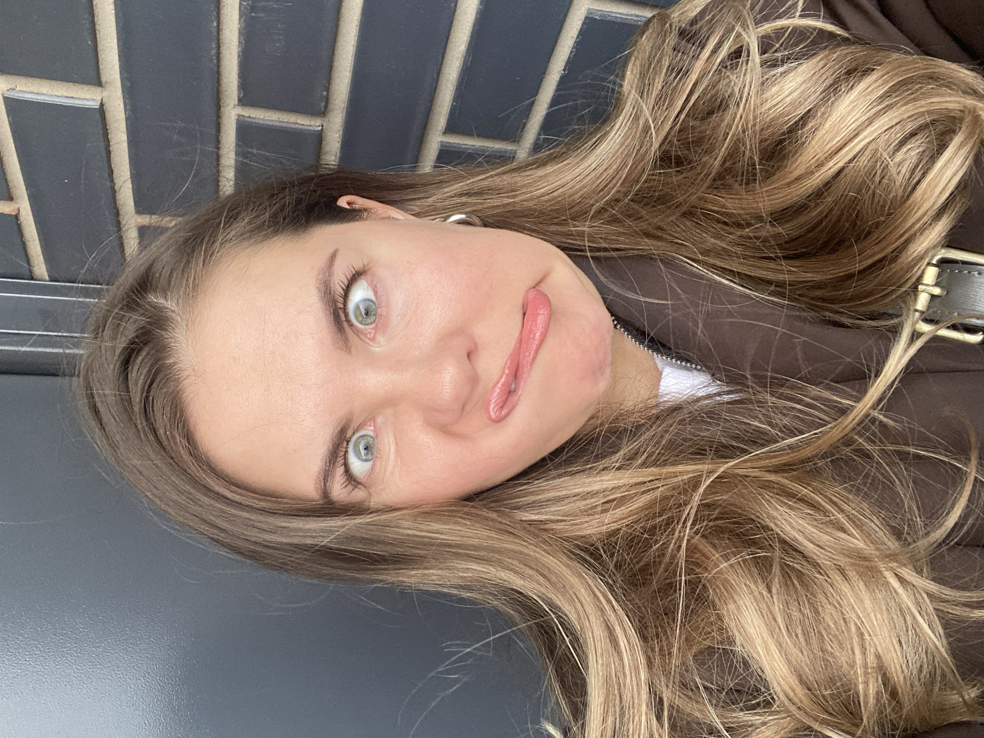 A young woman with light brown hair and green eyes is posing for a selfie. She is tilting her head to the side and sticking out her tongue, making a playful expression. She is wearing a brown jacket with a white shirt underneath. The background consists of dark gray brickwork and a gray surface. No information suggests a location in Izhevsk, Russia.