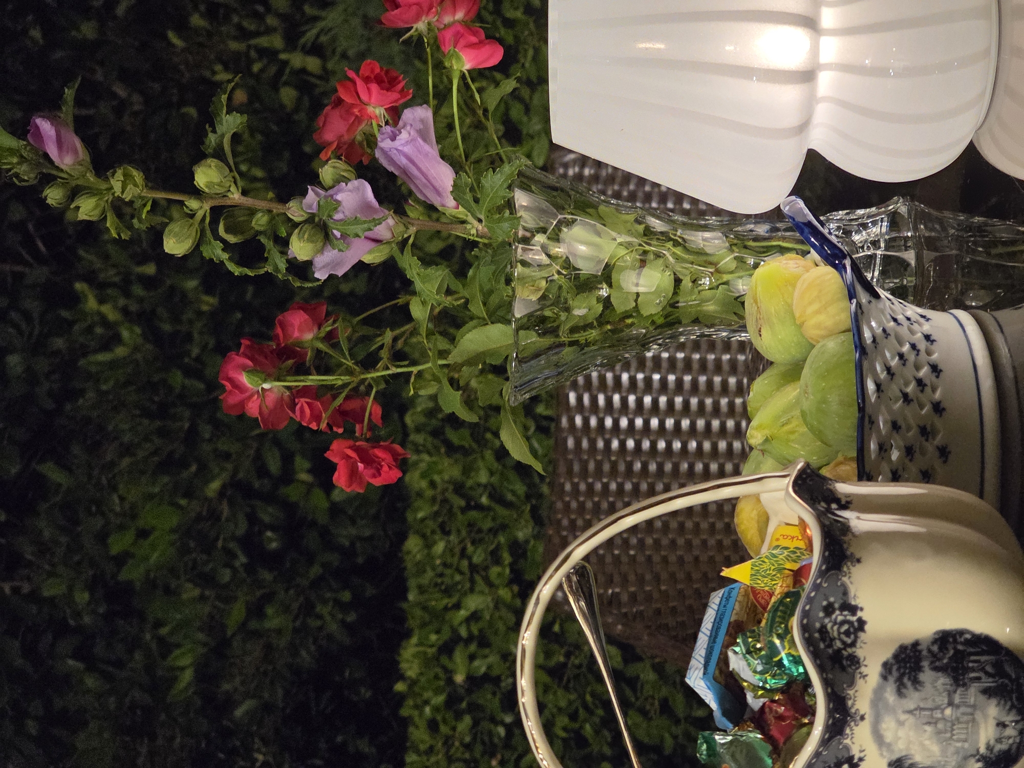 The image depicts an outdoor evening setting on a table. In the foreground, a decorative white and blue ceramic basket with a handle is filled with individually wrapped candies and what appears to be some small trinkets. To the right of the basket is a small blue and white patterned dish filled with green figs. Behind these, a clear glass vase holds a bouquet of red and pink flowers, interspersed with purple hibiscus blooms and green foliage. To the far right, a white, ribbed glass lantern is illuminated from within. The background shows a dark, manicured hedge. The setting appears to be a patio or garden area, possibly in Buzovna, Azerbaijan, given the context. No people are visible in the image.