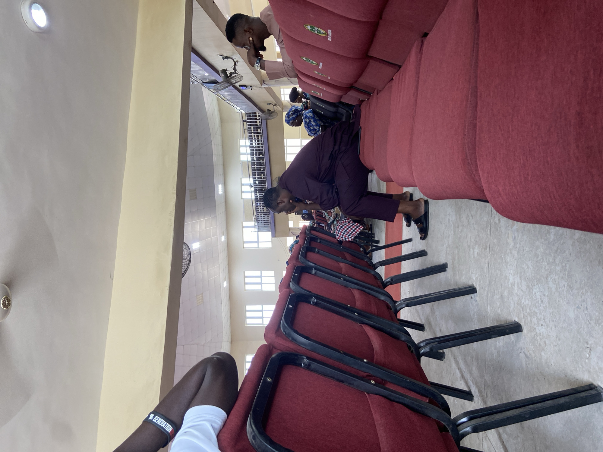 The image depicts an indoor scene within a large assembly hall located in Mararaba, Nigeria. The perspective is low, looking forward and slightly upward between rows of seating.
In the immediate foreground, on the left, a dark-skinned arm and bent knee of a person wearing a white short-sleeved top are visible. A black wristband with white text, which appears to read "GENERATION," is worn on the arm.
The hall is filled with rows of red seating. On the left side, individual red padded chairs with black metal frames are arranged. On the right side, continuous rows of red cushioned bench-style seating are present. The backs of these bench seats feature green and white logos or emblems, with some text visible, partially reading "SEAT OF GRACE."
Several individuals are seated. In the middle ground, on the left, a dark-skinned man wearing a dark purple traditional long-sleeved outfit and matching trousers is seated on the edge of a red cushion, facing left and slightly forward. His right elbow rests on his right knee, and his right hand is raised to his chin. He is wearing black slide sandals.
Further back and to the right, another dark-skinned man, wearing a light brown or pink traditional long-sleeved outfit, is seated in a similar posture, looking forward and slightly left. His left hand is near his mouth or chin, and he wears a silver-colored watch on his left wrist. He is also wearing dark slide sandals. Between these two men and behind the left man, several other people are partially visible, including a woman in a blue patterned headscarf.
The interior architecture features light-colored walls, with a lower section painted a pale yellow. A dark horizontal beam or ledge separates the wall sections. The ceiling is light-colored, featuring a mix of flat surfaces, square panels, recessed circular light fixtures, and flush-mounted rectangular lights. A decorative ceiling fan is partially visible in the upper left. In the upper right background, a dark metal railing indicates an elevated balcony or upper level, beneath which a large black wall-mounted oscillating fan is affixed. Multiple tall, narrow rectangular windows are visible high on the far wall, admitting natural light. The floor is a light grey, textured surface, appearing to be concrete.