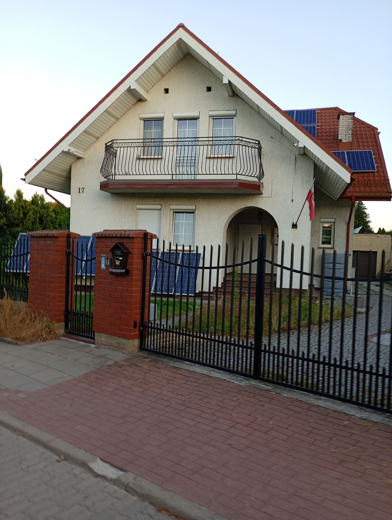 The image shows a residential house located at 17 in the city of Białystok, Poland. The house is a two-story structure with a light-colored facade and a red tiled roof. There are solar panels installed on the roof and on the ground in front of the house. A black metal fence surrounds the property, with a brick gatepost that has a mailbox attached. A Polish flag is displayed on the front of the house. The street in front of the house is paved with red bricks. The sky is clear and a light blue.