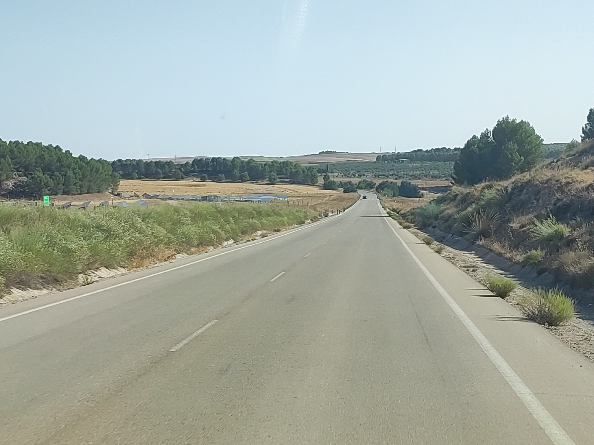 The image shows a wide, asphalt road stretching into the distance under a clear, pale blue sky. The road has white lane markings. On the left side of the road, there is a low embankment covered in green vegetation and some dry brush. Further to the left, there are some solar panels visible amidst a field of dry grass and scattered trees. On the right side of the road, the terrain rises into a dry, scrub-covered hillside with some scattered bushes casting shadows. In the distance, the road curves slightly, and a few trees and rolling hills are visible on the horizon, with some man-made structures and what appear to be wind turbines in the far distance. A single dark-colored car is visible on the road in the middle distance. The scene depicts a rural landscape in Huelves, Spain, during daylight.