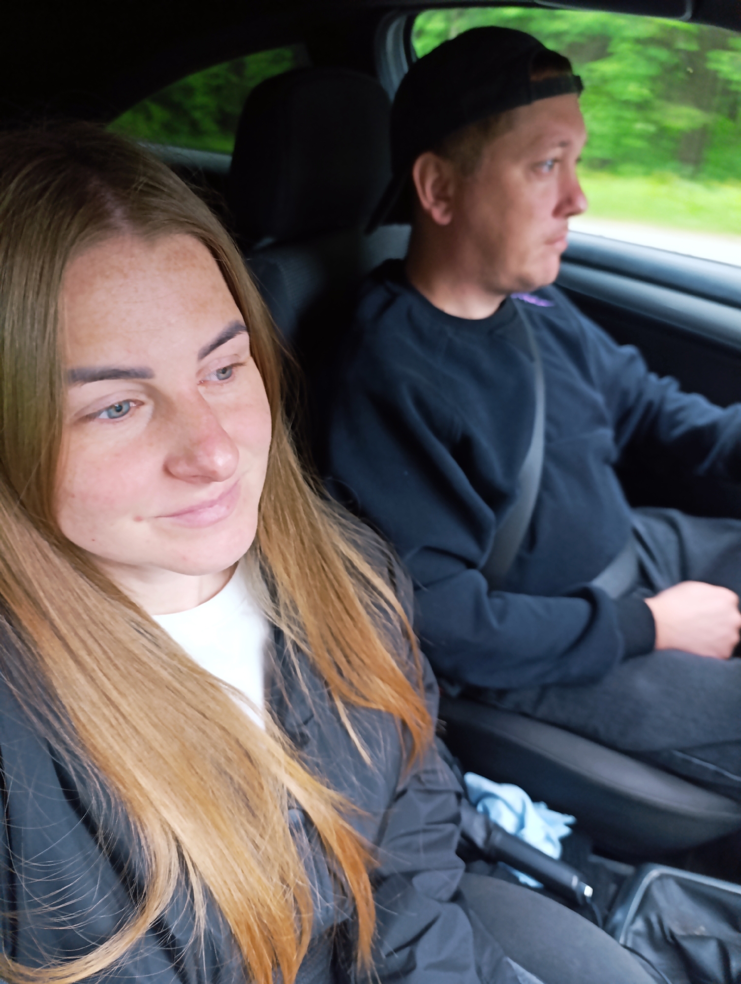 The image shows two people in a car, presumably in the Unknown, Unknown area. A woman with long, light brown hair and blue eyes is in the passenger seat, wearing a white shirt and a dark jacket. She is looking towards the front with a slight smile. The driver, a man wearing a black baseball cap and a dark blue sweatshirt, is looking towards the window, focused on driving. He has his seatbelt on. The interior of the car is visible, including the dashboard, seats, and a glimpse of the outside scenery through the windows. The light suggests it is daytime.