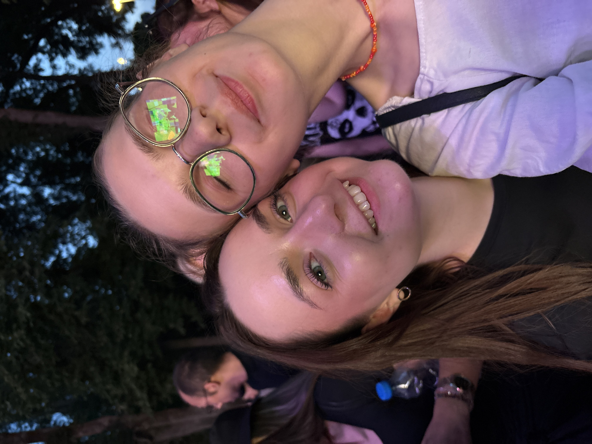 Two young women pose closely for a selfie. The woman on top wears round glasses with a complex reflection in the lenses. She has a beaded necklace. The woman below smiles, showing her teeth, and has brown hair. In the background are trees, the sky, and indistinct people. There is a water bottle visible. While I cannot definitively confirm the exact location in Kragujevac without additional information, the setting suggests an outdoor public space.