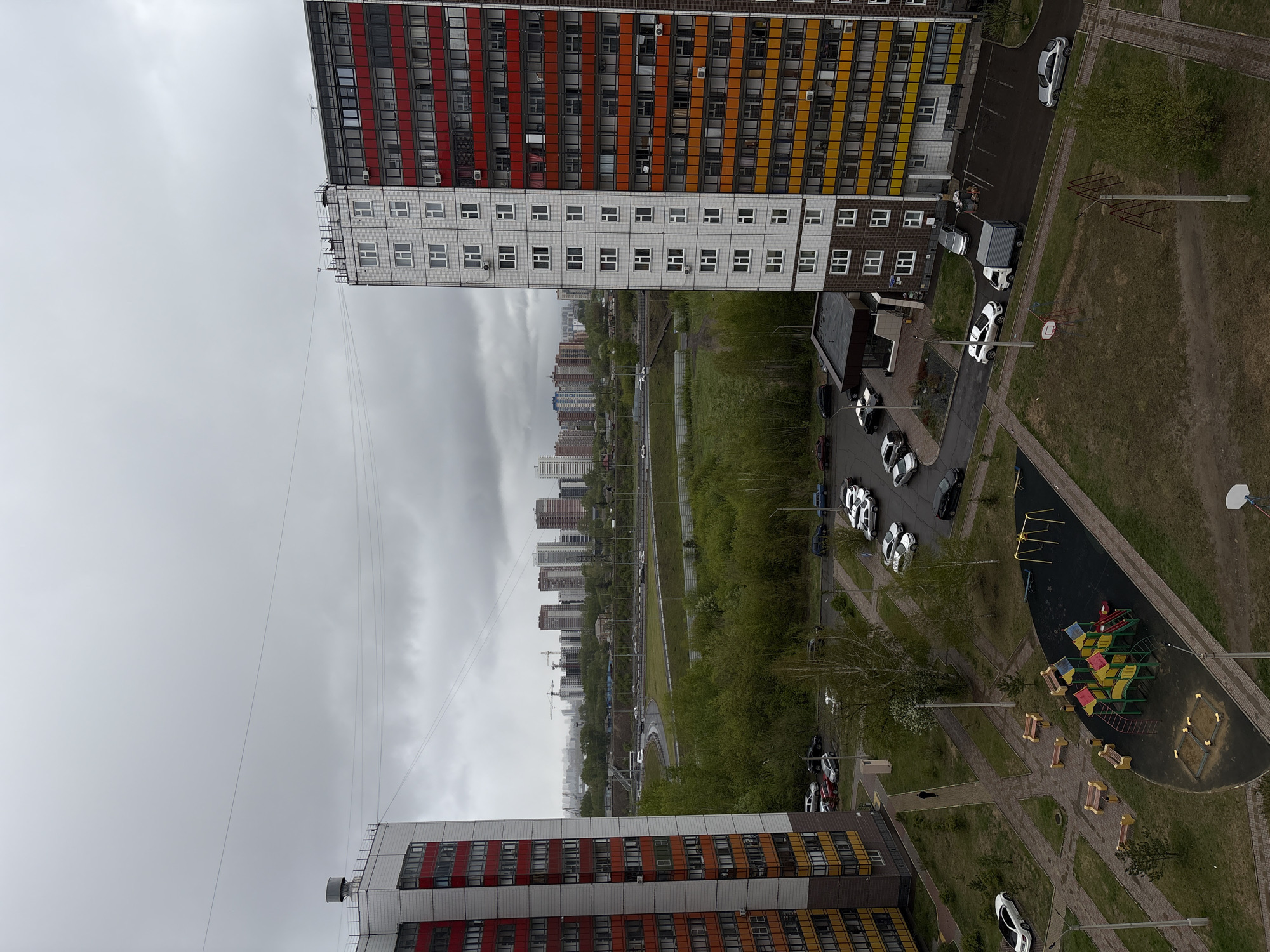 This is an elevated, tilted view of a cityscape, possibly Krasnoyarsk, Russia. The foreground features two high-rise apartment buildings with distinctive horizontal stripes of red, orange, and yellow. The buildings have numerous windows, indicating a residential area.
Between the buildings lies a green space containing trees, grass, a playground with colorful equipment, and a network of paved walkways. Several cars are parked on the streets adjacent to the buildings.
In the distance, a row of more high-rise buildings is visible under a cloudy sky. There are also construction cranes, further indicating urban development. A road runs parallel to the row of buildings.
The scene appears to be taken during an overcast day.