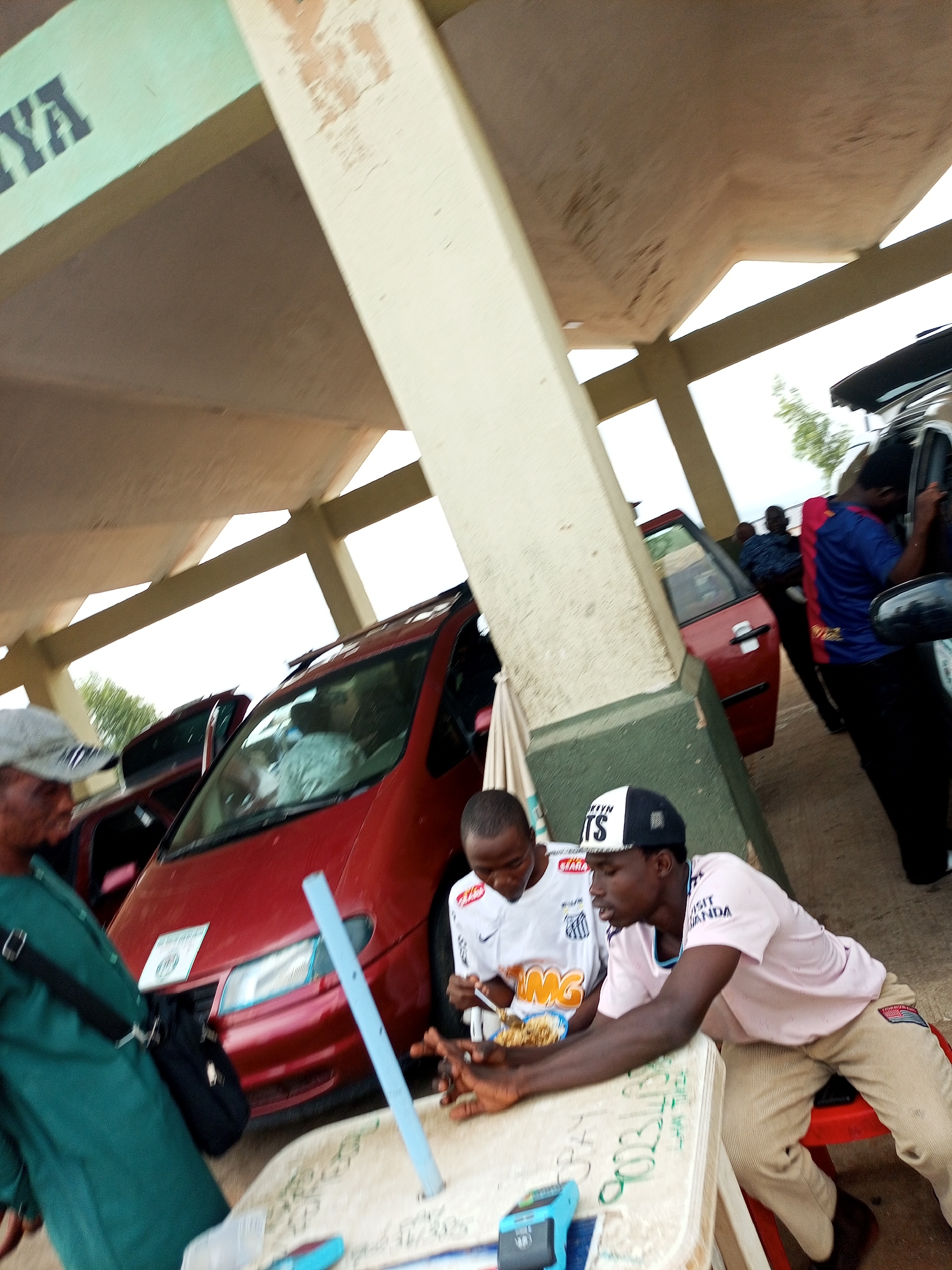 In Gombe, Nigeria, the image depicts an outdoor scene under a covered structure. Two young men are seated at a table, with one man in a white t-shirt eating from a bowl. Another man in a light pink t-shirt and tan pants is leaning on the table, looking at his phone. A third man in a green garment stands to the left, partially obscured. A red car is parked behind the table. The environment appears to be a public space with a concrete floor and support beams.