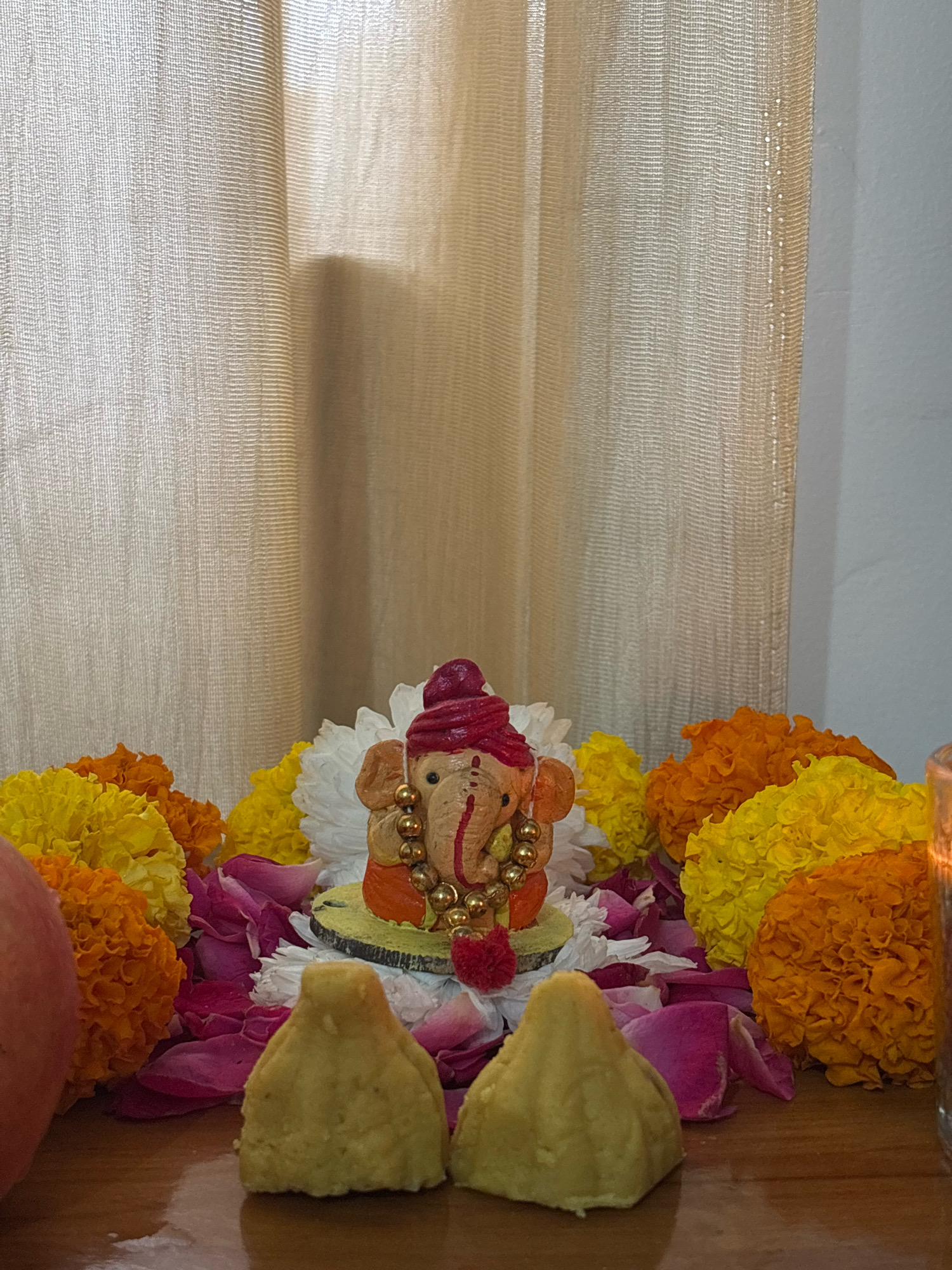 A statue of the Hindu deity Ganesha is placed on a wooden surface, surrounded by colorful marigolds, rose petals, and two modaks. Ganesha is depicted with a red turban and gold jewelry. The scene is lit by natural light filtering through a sheer beige curtain in the background. The image appears to be taken indoors. There is no information in the image to suggest a location in Bhatta, India.