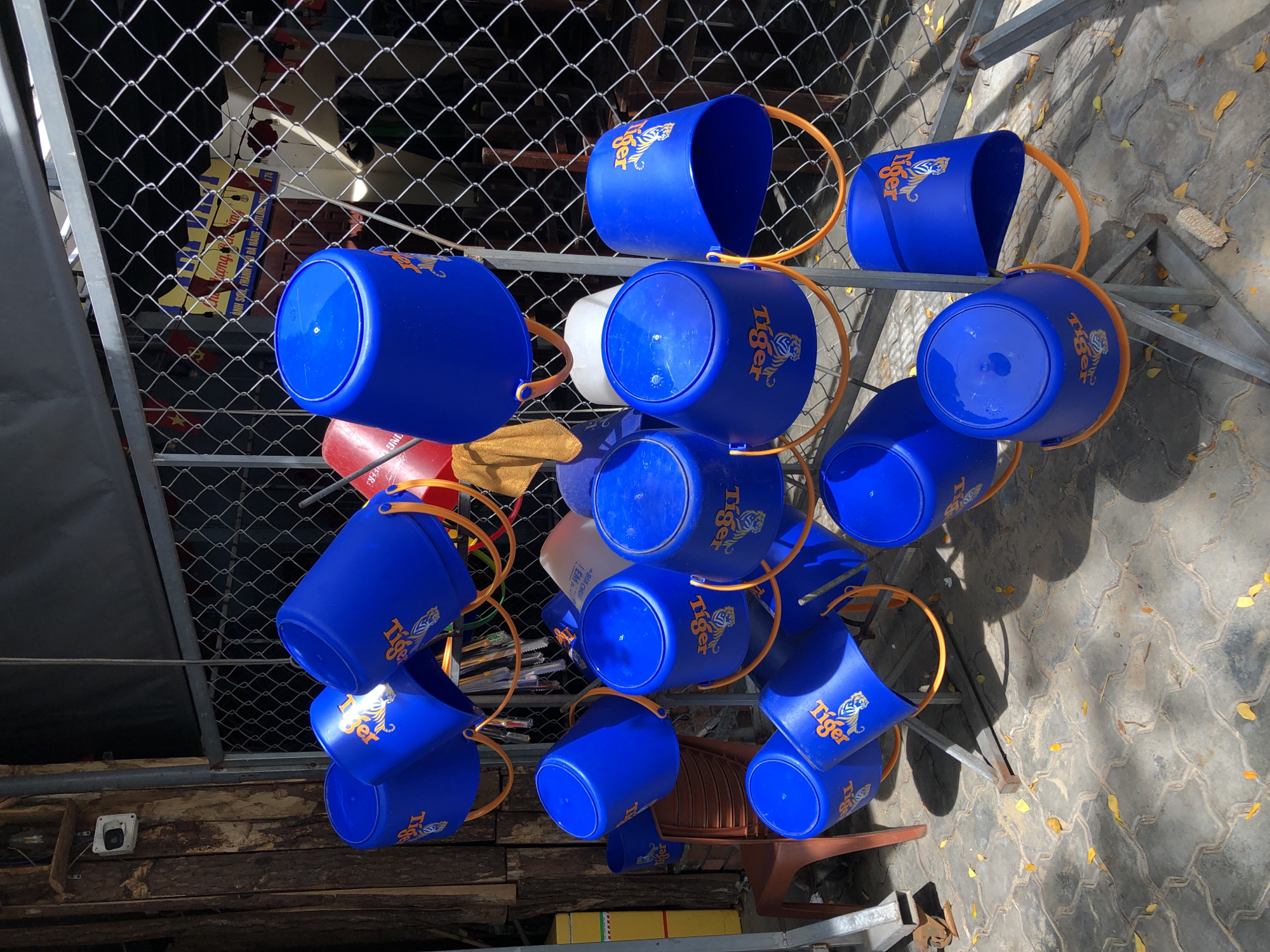 An outdoor scene in Đà Nẵng, Vietnam, features a rack of bright blue plastic buckets, many emblazoned with the orange "Tiger" logo, arranged in a staggered, hanging formation. The buckets are of various sizes and orientations, with some facing upwards and others downwards, all suspended by orange handles. The rack itself appears to be constructed from gray metal poles. Behind the buckets, a wire mesh fence encloses an area with some vegetation visible. To the left of the buckets, a rough-hewn wooden post and a brown plastic chair are present. The ground is paved with interlocking gray pavers, sparsely scattered with fallen yellow and orange leaves, suggesting an autumn setting or a tree in close proximity. The lighting indicates a bright, sunny day, casting shadows from the buckets and the rack onto the ground. The overall impression is one of an outdoor storage or display area, possibly for a business, given the branded buckets.
