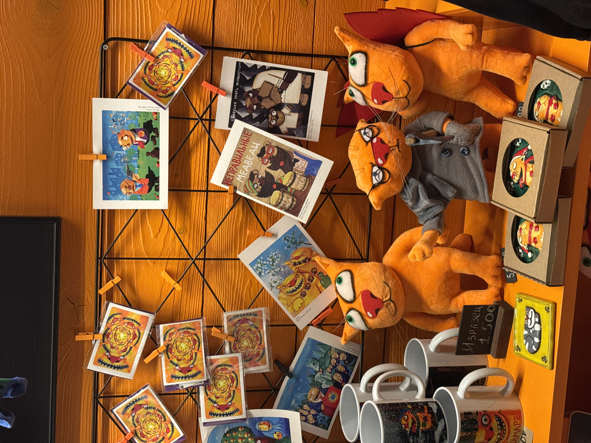 An indoor, eye-level shot shows a display of merchandise, likely in a store. The merchandise includes several orange cat plush toys, mugs with cartoonish designs, and small framed prints or postcards. Three plush cats are prominently displayed: one in the center wearing a grey coat and glasses, and two on the right, one with red wings. The cats are positioned on an orange surface, in front of a black wire grid attached to an orange wooden wall. Several cards with colorful, mandala-like patterns and some with cartoon illustrations are clipped to the wire grid. Two of the illustrated cards feature bears and text in Russian, with one reading "НЕПРАВИЛЬНЫЕ МЕДВЕДИ" (Incorrect Bears) and the other "БАБУШКЕ ЗНАТЬ НЕОБЯЗАТЕЛЬНО" (Grandmother Doesn't Need to Know). A small blackboard sign with "Изразец 1.500" written on it stands on the orange surface. Several mugs, some stacked, are visible to the left of the cats, with one mug featuring a cartoon character. In the foreground, on the orange surface, are two small cardboard boxes, each containing a round, cookie-like object with a cartoon image of a cat. Another yellow square tile with a black and white cat design is also visible. The lighting is even, suggesting an interior setting. The overall scene is cluttered but organized, showcasing various items for sale.