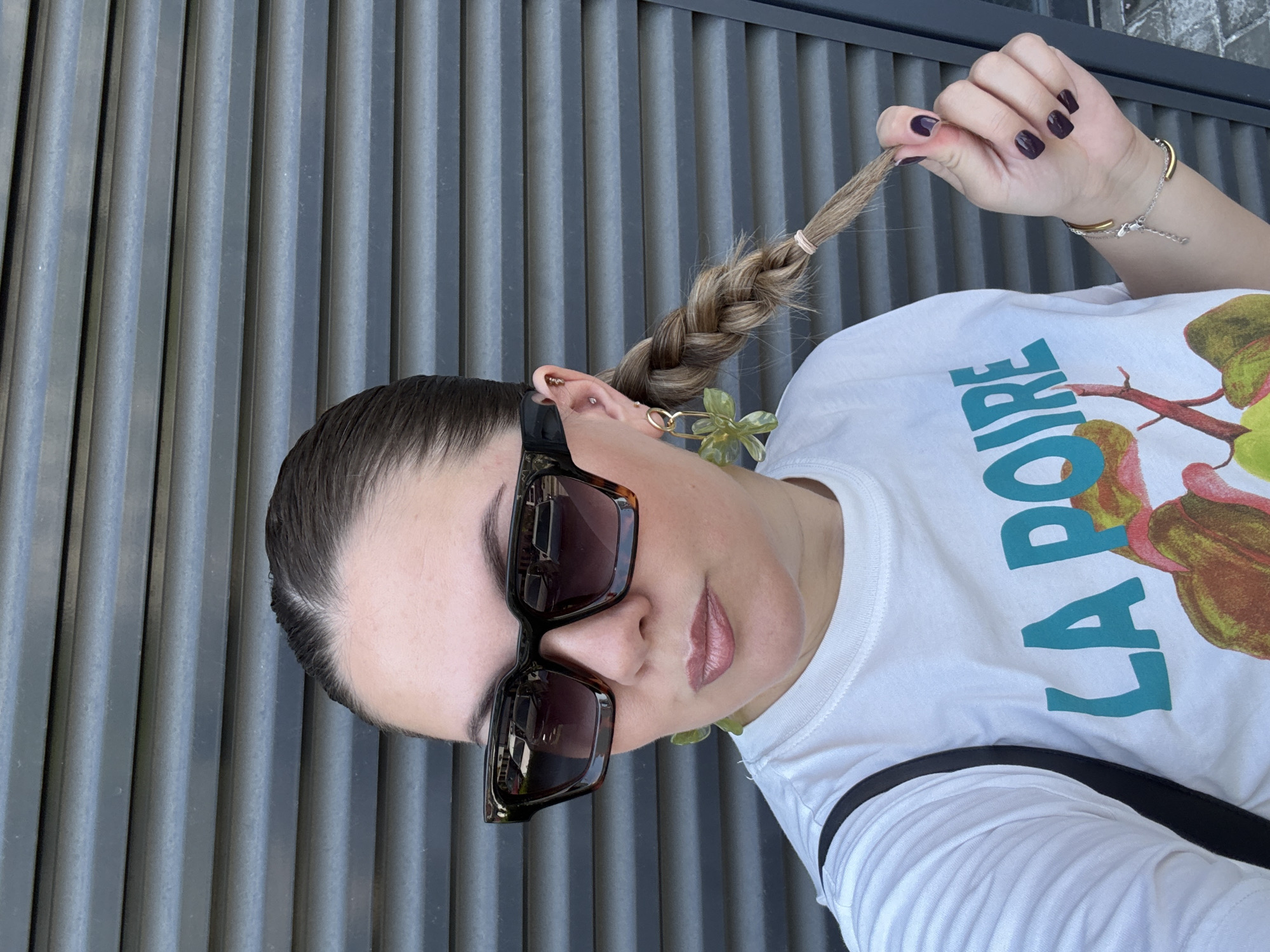 A fair-skinned woman with dark brown hair braided to the side is holding the end of her braid in her hand, showing a dark purple manicure. She is wearing sunglasses, a white t-shirt with the text "LA POIRE" and a floral design. The location is Izhevsk, Russia, based on the text in the image. The background is a gray corrugated wall.