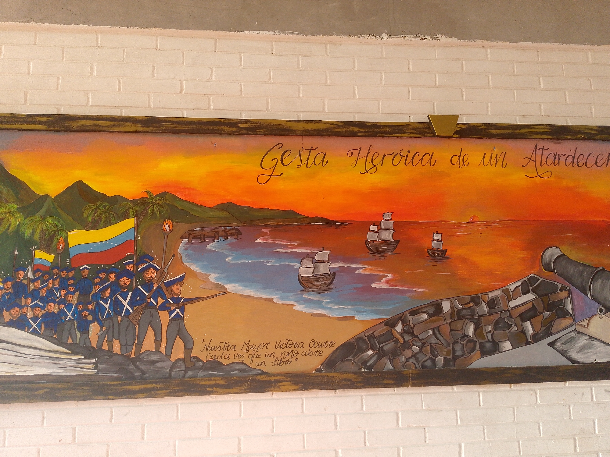 The image shows a rectangular mural painted on a white brick wall. The mural depicts a coastal scene, likely representing a historical event. A group of soldiers in blue uniforms with white crosses on their chests is marching along a sandy beach, carrying the flag of Venezuela. In the background, there are three sailing ships on the water under an orange and yellow sunset. To the right, there is a depiction of a stone wall with a cannon. The title "Gesta Heroica de un Atardecer" is written above the sunset sky. Beneath the soldiers are the words "Nuestra Mayor Victoria Ocurre Cada vez que un niño abre un Libro." The scene is likely intended to represent a battle or heroic event that occurred in Juan Griego, Venezuela, where historical sites often depict naval battles.