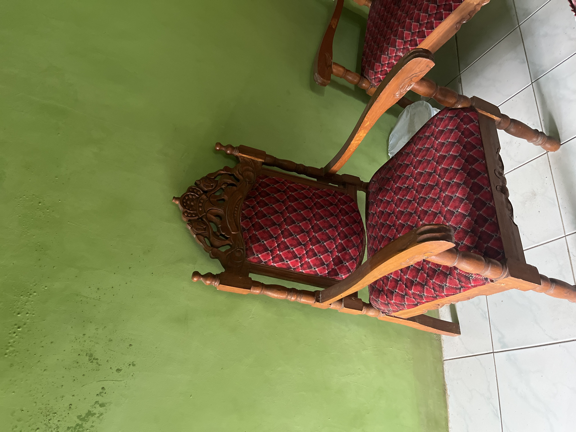 In a room with a textured, bright green wall, two ornate wooden chairs with red patterned upholstery are positioned on a white tiled floor. The chair in the foreground is facing slightly towards the viewer, showcasing its carved wooden backrest, turned wooden legs, and arms. The second chair is partially visible to the right, in the background, and appears to be of similar design. A white sheet or cloth is visible on the floor between the two chairs. The lighting suggests it is daytime, with shadows cast by the chairs. The location is identified as Glagah Kulon, Indonesia.