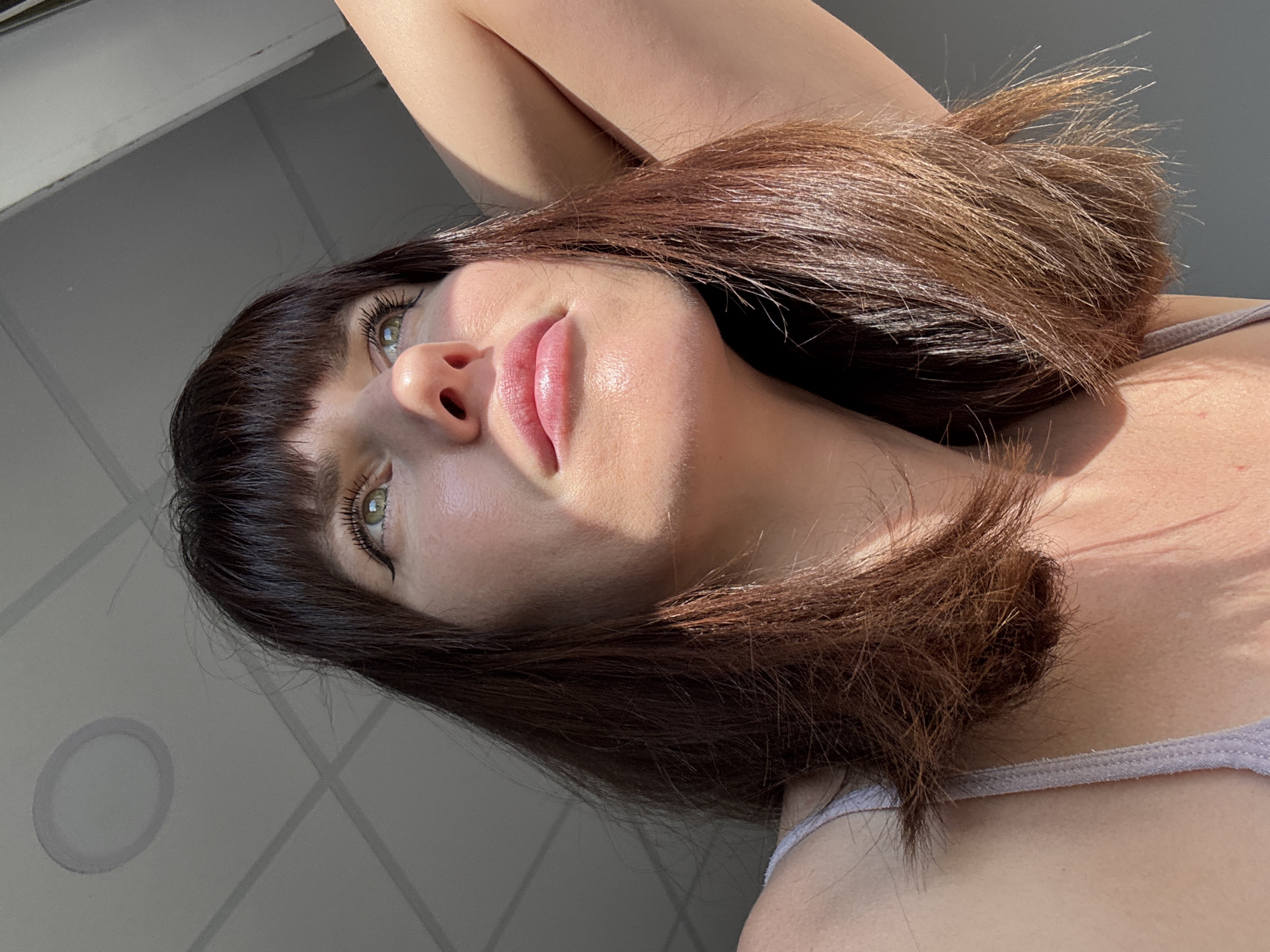 A young woman with dark brown hair and bangs, wearing a light-colored tank top, is captured in a close-up shot looking upwards and to her right. Her eyes are light green, and she is wearing eyeliner. Sunlight is falling on her face, highlighting her features, including her lips which appear to be glossy. Her right arm is raised and bent, with her hand resting behind her head. The background shows a ceiling with visible tiles and a recessed light fixture on the left, and a window frame on the right. The environment appears to be indoors, possibly in an apartment or office. The image is taken from a low angle, looking up at the woman.