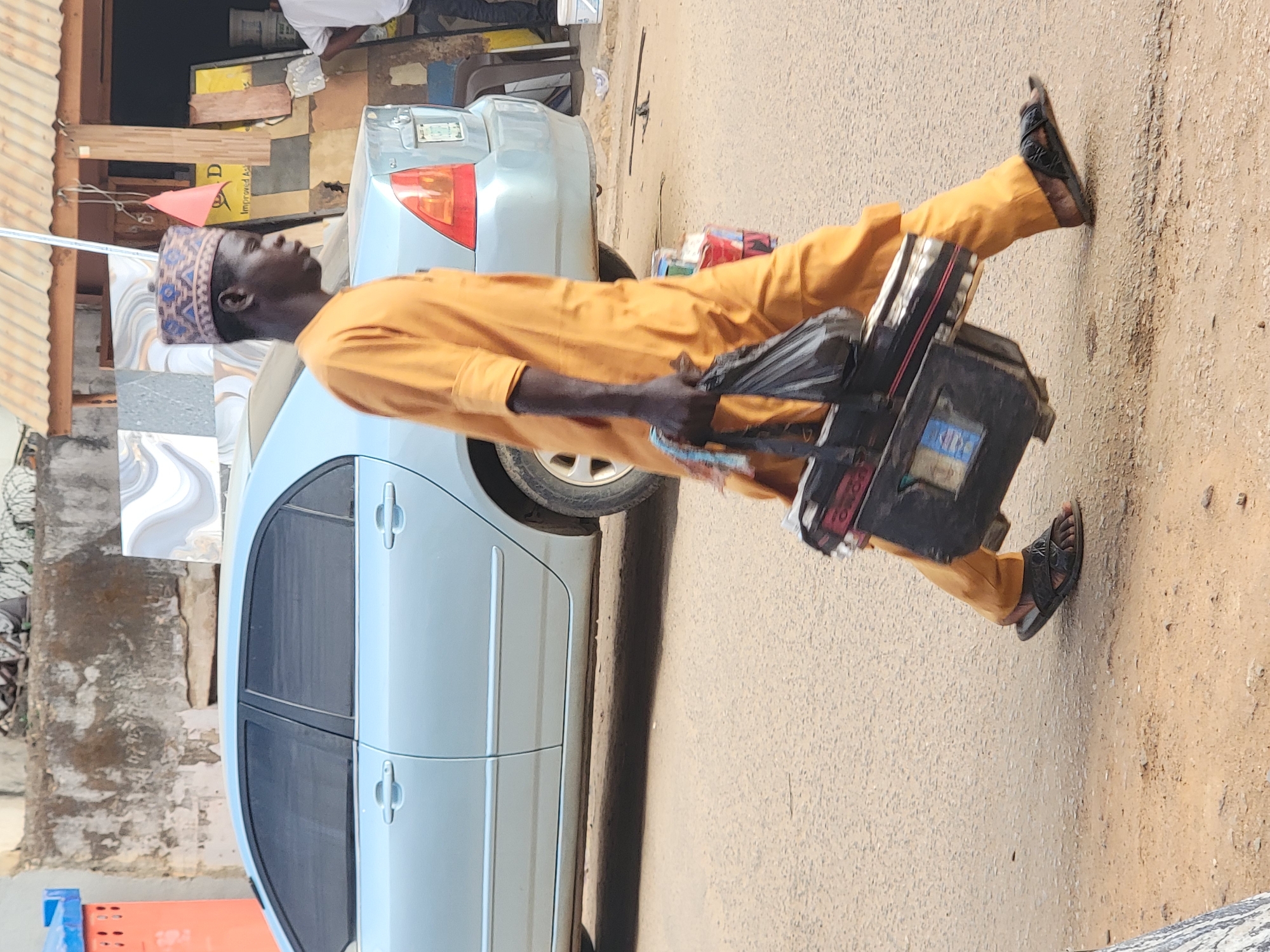 A man, presumably in Maiburiji, Nigeria, is walking on a paved street next to a parked light blue car. He is wearing a light orange outfit with a patterned blue and white kufi cap and sandals. He carries a black toolbox. The scene appears to be a street or roadside in an urban setting.