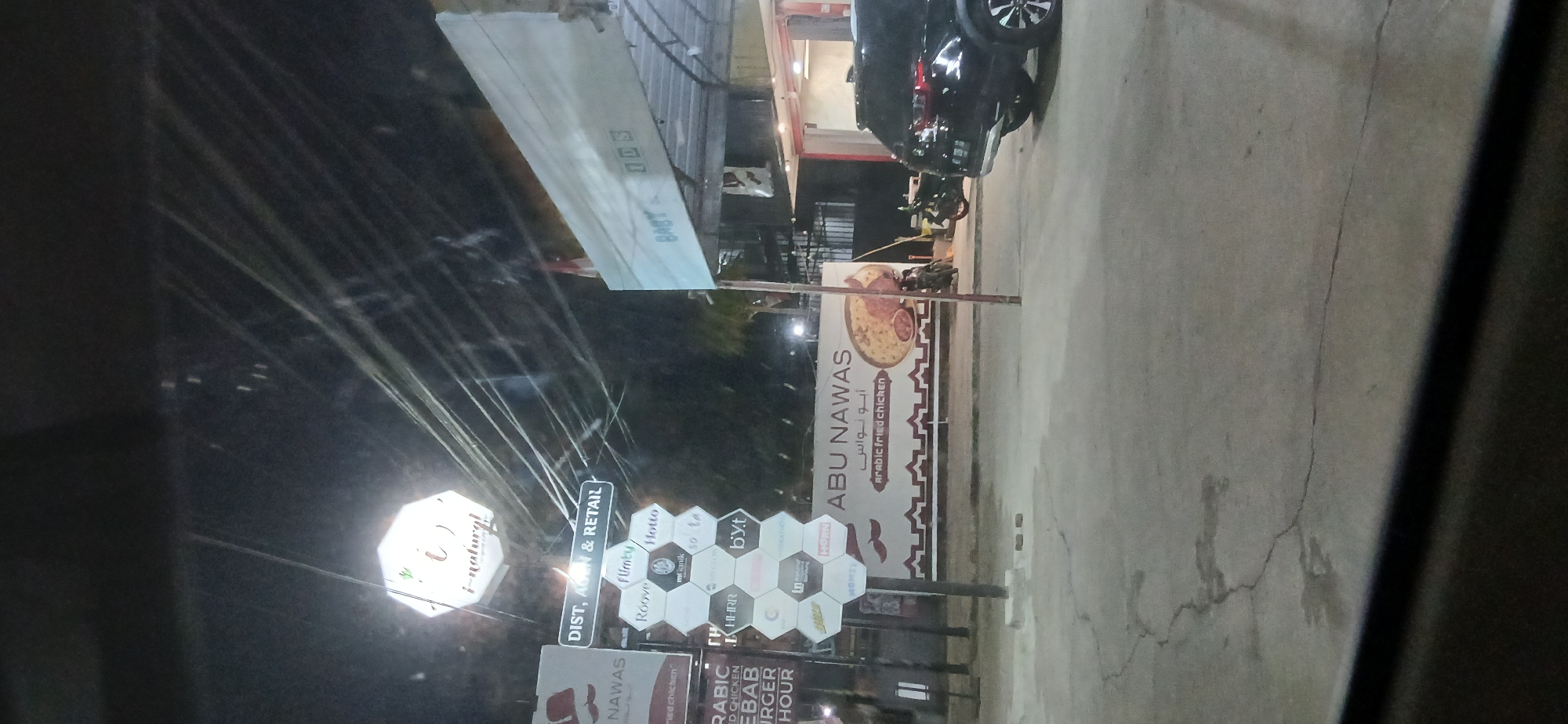 The image depicts a night scene in Samarinda, Indonesia, likely near a commercial establishment. In the foreground, a cracked concrete surface is visible, partially obscured by a dark, reflective surface that could be a car window. A black SUV is parked to the right, with its rear end facing the viewer.
Behind the car, a large white banner with the text "ABU NAWAS" and Arabic script is displayed, advertising "ARABIC FRIED CHICKEN." To the left of the banner, a hexagonal display board features various logos, including "fluby," "Roove," "Hotte," "mtanik," "so ta," "HYHRR," "byt," "C," "In," and "HORN," suggesting a commercial area with multiple businesses.
Above the hexagonal display, a directional sign reads "DIST, AGEN & RETAIL." Further to the left, a sign with a stylized mustache and the name "NAWAS" is partially visible. A prominent octagonal sign with a leaf logo and the text "i-natural" illuminates the scene. Overhead, a tangle of electrical wires crisscrosses the night sky. Two motorcycles are parked near the banner. The overall impression is a bustling street scene at night, illuminated by artificial lights and signage.