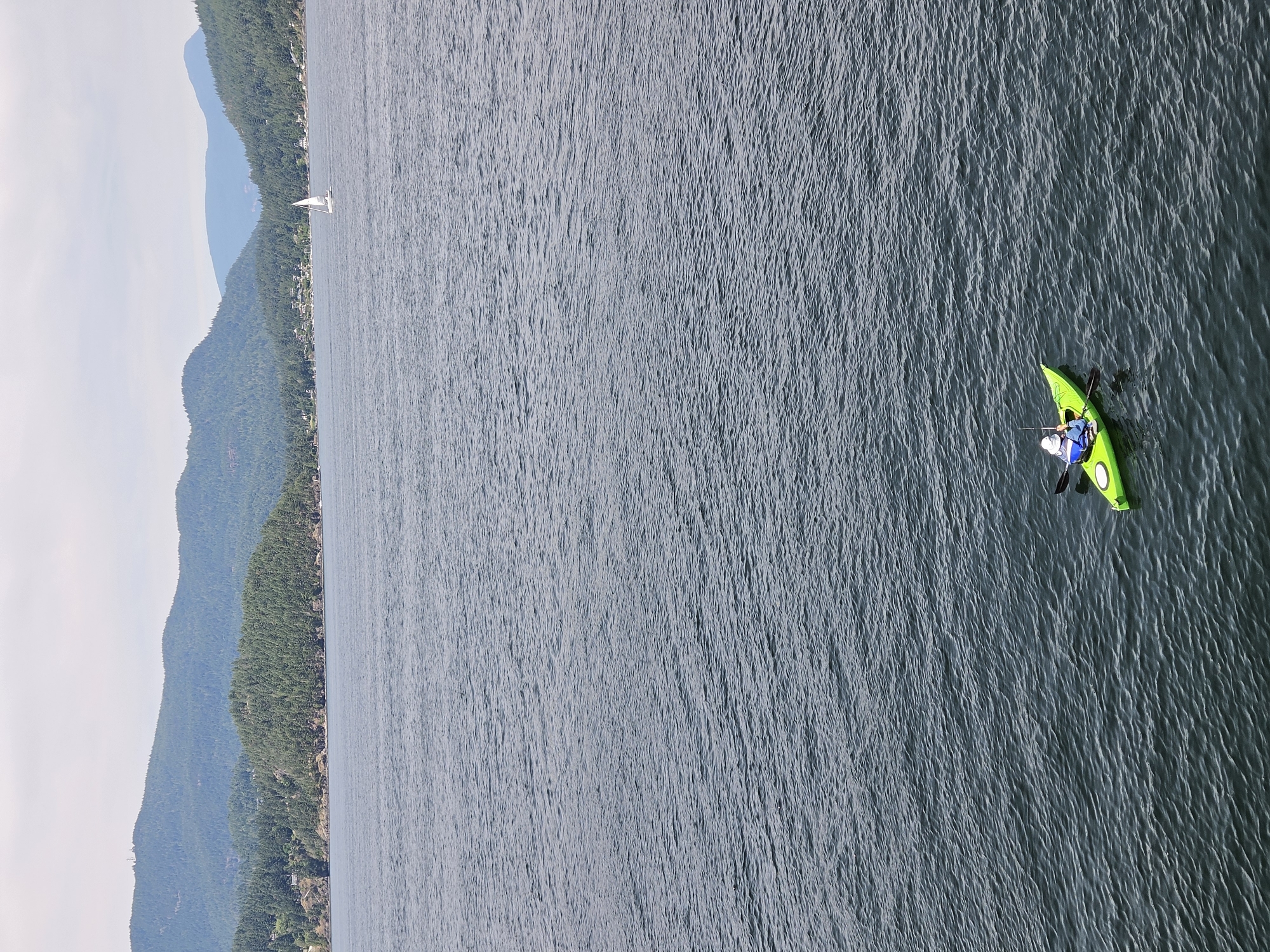 This media file captures an aerial view of a person kayaking on a body of water, likely in West Vancouver, Canada, given the context. The scene is dominated by the dark, rippling surface of the water, which occupies most of the frame. To the left, a lush, tree-covered mountainside slopes down towards the water's edge, with scattered houses visible along the shore. Further in the distance, hazy blue mountains form a layered backdrop against a pale, overcast sky, suggesting a cloudy or misty day.
In the foreground, on the right side of the image, a person is actively paddling a bright green kayak. The kayaker is wearing a blue shirt and a white helmet, and they are holding a black paddle, propelling themselves through the water. The kayak has visible yellow accents. The water around the kayak shows small ripples, indicating movement. In the distance, a solitary white sailboat can be seen near the shore, adding another element to the aquatic scene. The overall impression is one of a peaceful, outdoor activity set against a scenic natural landscape. There is no visible text in the media file.