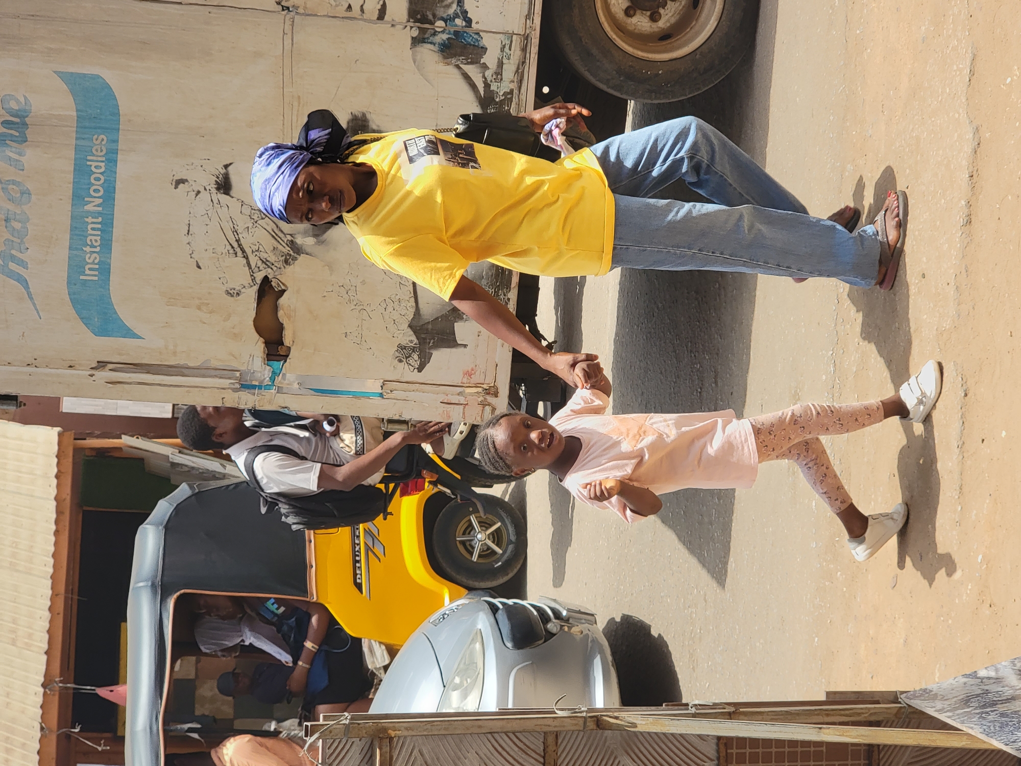 A woman in a yellow t-shirt and blue jeans holds the hand of a young girl wearing a light pink shirt and patterned leggings in what appears to be a street scene in Maiburiji, Nigeria. The woman is standing next to a white truck with "Instant Noodles" printed on the side. A yellow "Deluxe" tricycle (auto rickshaw) is parked nearby, with a person wearing a white shirt and a black backpack standing beside it. Another person is visible inside the tricycle. A silver car is parked in front of the tricycle. The ground is a mixture of asphalt and dirt. The scene suggests a busy urban environment.