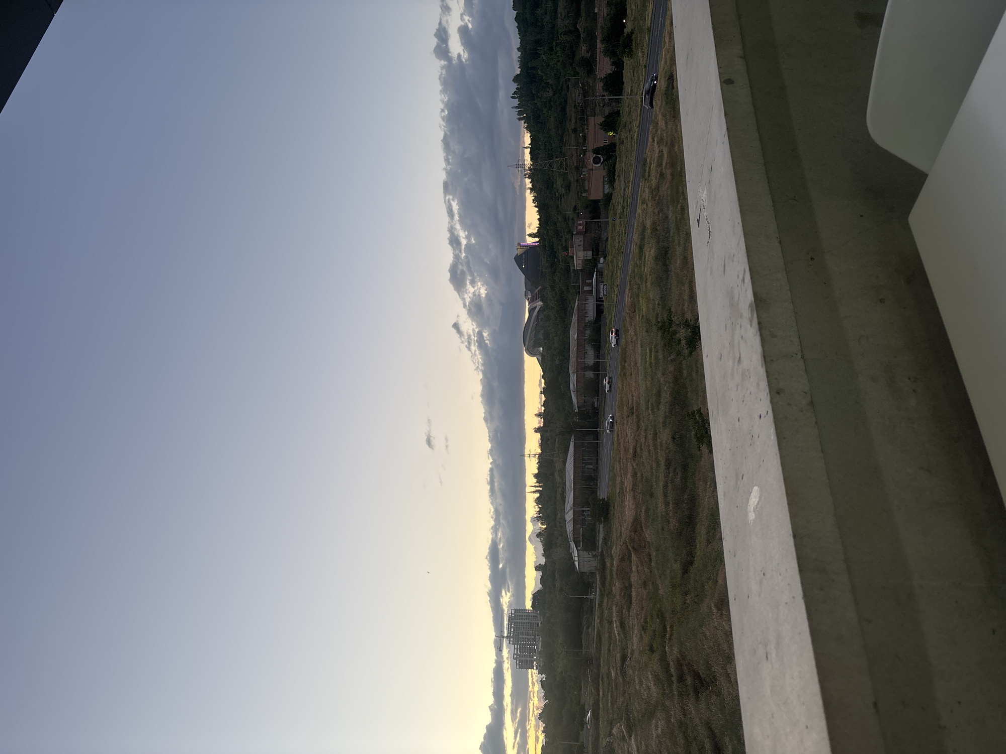 The image presents a wide, horizontally oriented landscape captured from an elevated perspective, likely a balcony or high-rise building, during either sunrise or sunset. The sky dominates the upper half, featuring a soft gradient from a warm, golden-yellow glow near the horizon to a pale blue and then a darker, muted blue-grey towards the top. A band of scattered, elongated clouds, some appearing dark, stretches across the sky, particularly in the mid-left section, illuminated from below by the low-angle sun.
The horizon line bisects the image horizontally, revealing a blend of natural and urban environments. Extensive fields of dry, brownish-green grass and patches of dense green trees, possibly indicating distant hills or mountains, form part of the landscape. A road runs through the scene, roughly parallel to the horizon, with at least two vehicles (one light-colored, one dark) visible on it. Various buildings, ranging from what appear to be residential structures to larger commercial or industrial types, are scattered across the mid-ground. A prominent construction site with a partially erected high-rise building and a crane is visible towards the lower left. An electricity transmission tower stands out against the horizon. A distinctive building with a rounded roofline and some internal lighting is also visible in the central-right mid-ground.
In the immediate foreground, a robust concrete railing or wall runs along the right edge of the frame, emphasizing the high vantage point. A section of a white, curved structure is also visible to the right of the concrete, possibly part of the building's architecture or outdoor furniture. The soft, warm light and long shadows indicate either the early morning or late evening. No specific human activity is the focus, and no text is visible within the image. The scene conveys a sense of calm and expansive view over an urban and natural landscape.