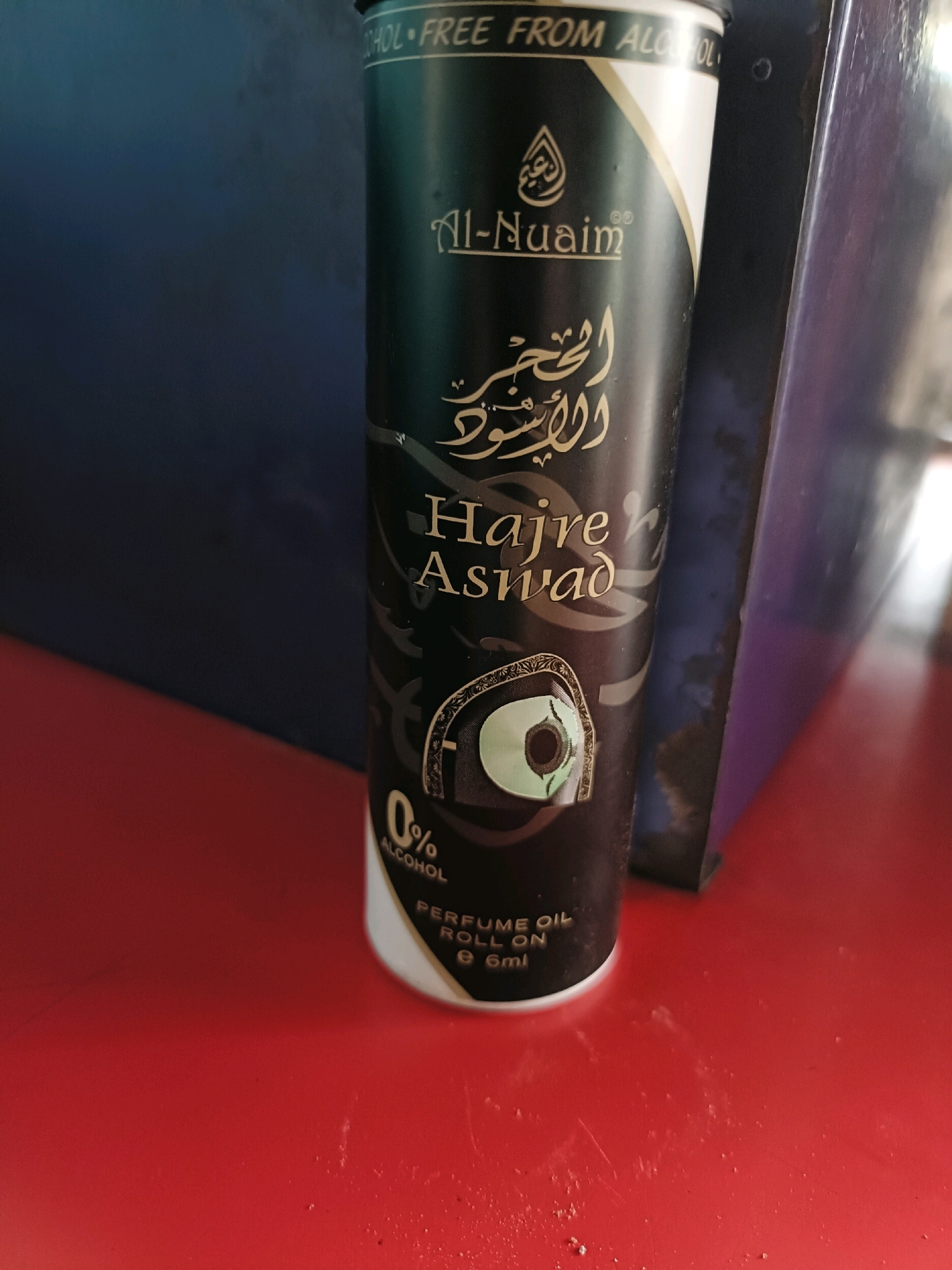 The image shows a close-up of a black cylindrical bottle of "Al-Nuaim Hajre Aswad" perfume oil. The bottle features gold lettering and an intricate design. A logo with a droplet shape is visible at the top. Below the brand name, Arabic script is present. The text "HAJRE ASWAD" is clearly legible in English. A decorative emblem with a green, circular element resembling an eye is featured on the bottle. Towards the bottom, "0% ALCOHOL" is prominently displayed, followed by "PERFUME OIL ROLL ON" and "e 6ml". The bottle is positioned on a red surface, with a dark blue and purple object partially visible in the background. The lighting suggests an indoor setting, possibly a retail environment or a personal collection. There is no indication of the city of Girjam, India, in the image.