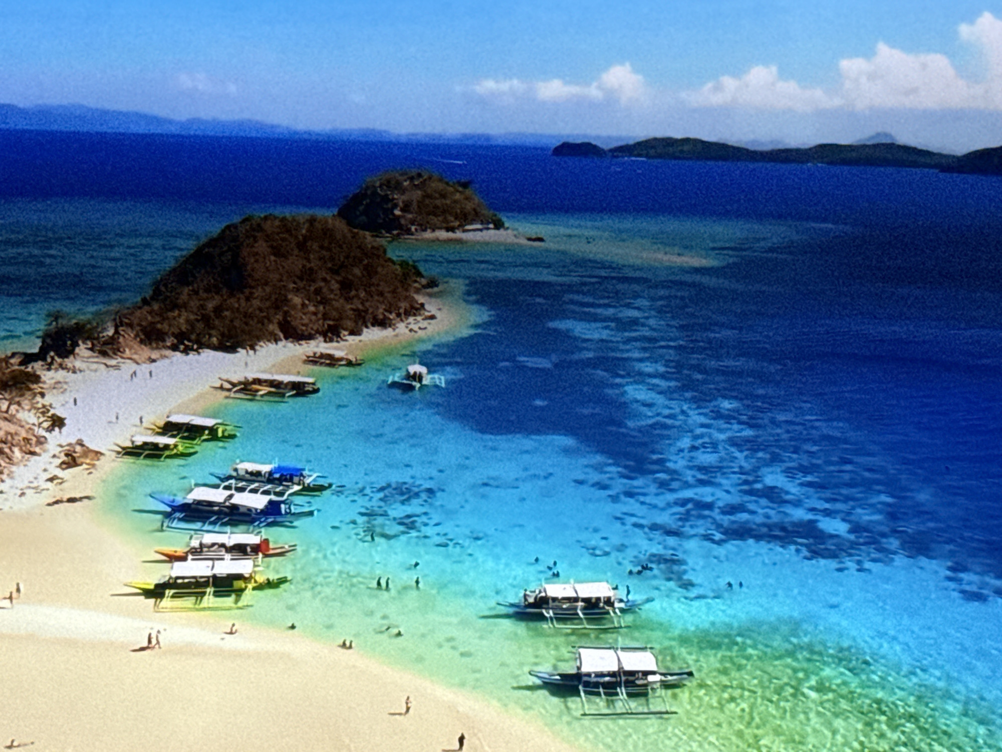 This is an aerial drone shot of a tropical beach scene with clear blue water and several boats anchored near the shore. The beach is sandy and sparsely populated with a few individuals walking along the coastline or swimming in the shallow waters. The boats are a mixture of traditional outrigger boats, some white with roofs and others with yellow hulls. In the background, there are several small, rocky islands covered in vegetation, and a larger landmass with mountains in the distance. The sky is a bright blue with a few wispy clouds. The scene depicts a picturesque and serene vacation spot, likely a popular tourist destination. There is no information to suggest this location is in Burgas, Bulgaria.