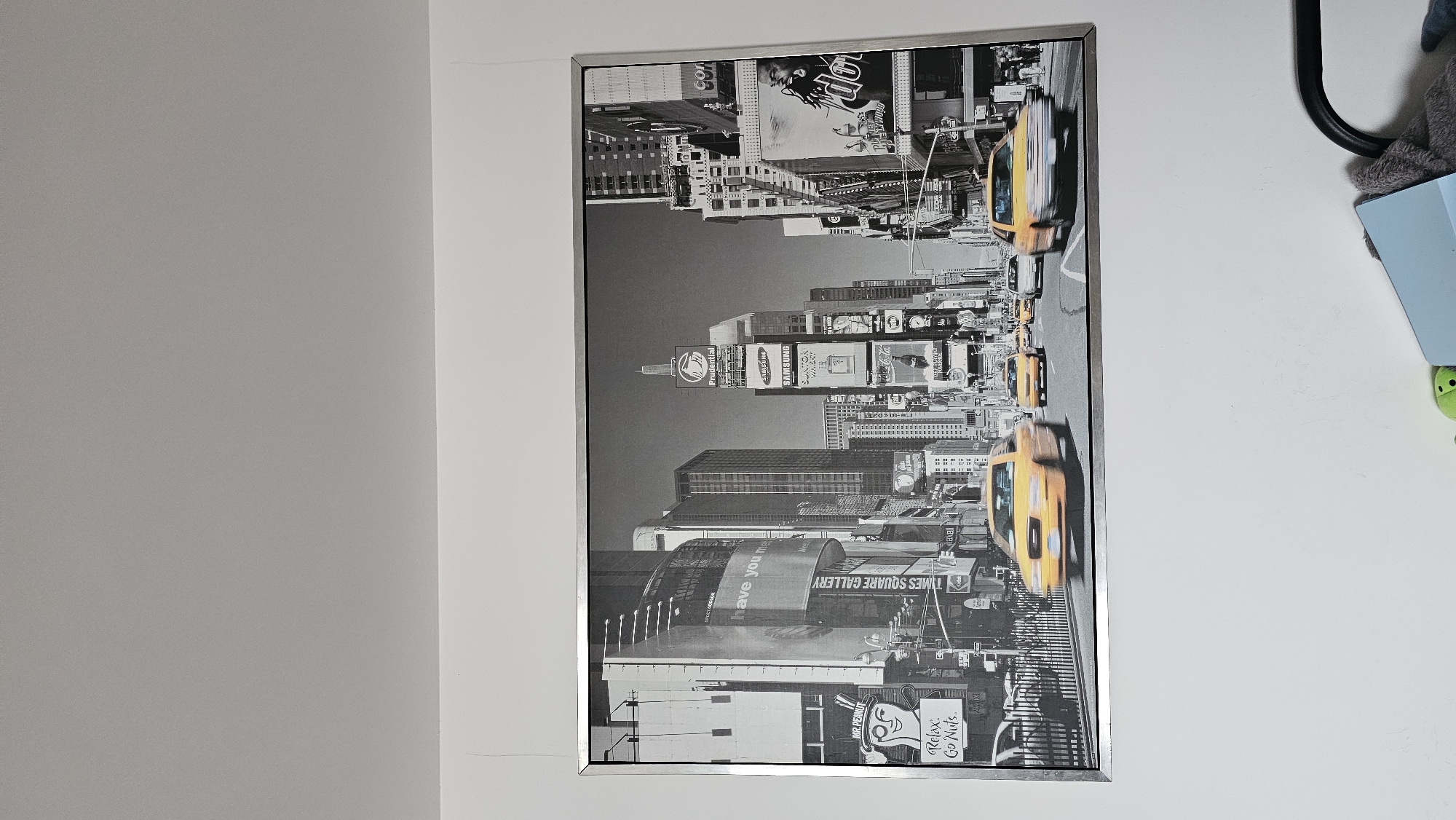 The primary image displays a large, rectangular framed artwork mounted on a plain white wall, positioned in a room possibly located in Boussu-lez-Walcourt, Belgium. The artwork itself is a black and white photograph depicting a dynamic urban street scene, clearly identifiable as Times Square, New York City. Numerous multi-story buildings dominate the background, densely covered with large advertising billboards and digital screens. Recognizable brand names and text visible on these displays include "Prudential," "Samsung," "Suntory Whisky," "Coca-Cola," "Mr. Peanut" accompanied by the slogan "Relax. Go Nuts.," and "TIMES SQUARE GALLERY." A street, separated by a dark fence, traverses the lower section of the photographic print. Multiple yellow taxi cabs are visible on the street, with several rendered in motion blur, suggesting active traffic and a busy environment. The frame enclosing the artwork is a metallic silver tone. Above the framed piece, the wall transitions to a light gray color, with a faint ceiling line visible at the top. The wall area below the artwork maintains the white color seen to its left and right. In the bottom right corner of the original image, a partial view of a black, possibly tubular, furniture element is present, along with indistinct blue and green objects resting on the floor.