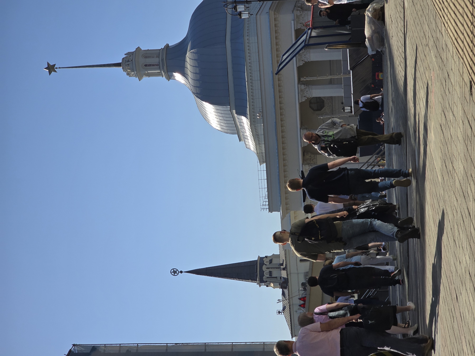 The image shows a daytime scene of people walking on a brick pavement in Moscow, Russia. In the background are architectural structures, including a building with a large, rounded blue dome and another with a tall spire topped with a star. The sky is clear and blue. The photograph appears to be taken at an angle. The location is likely near the Kremlin and Red Square, given the recognizable architecture.