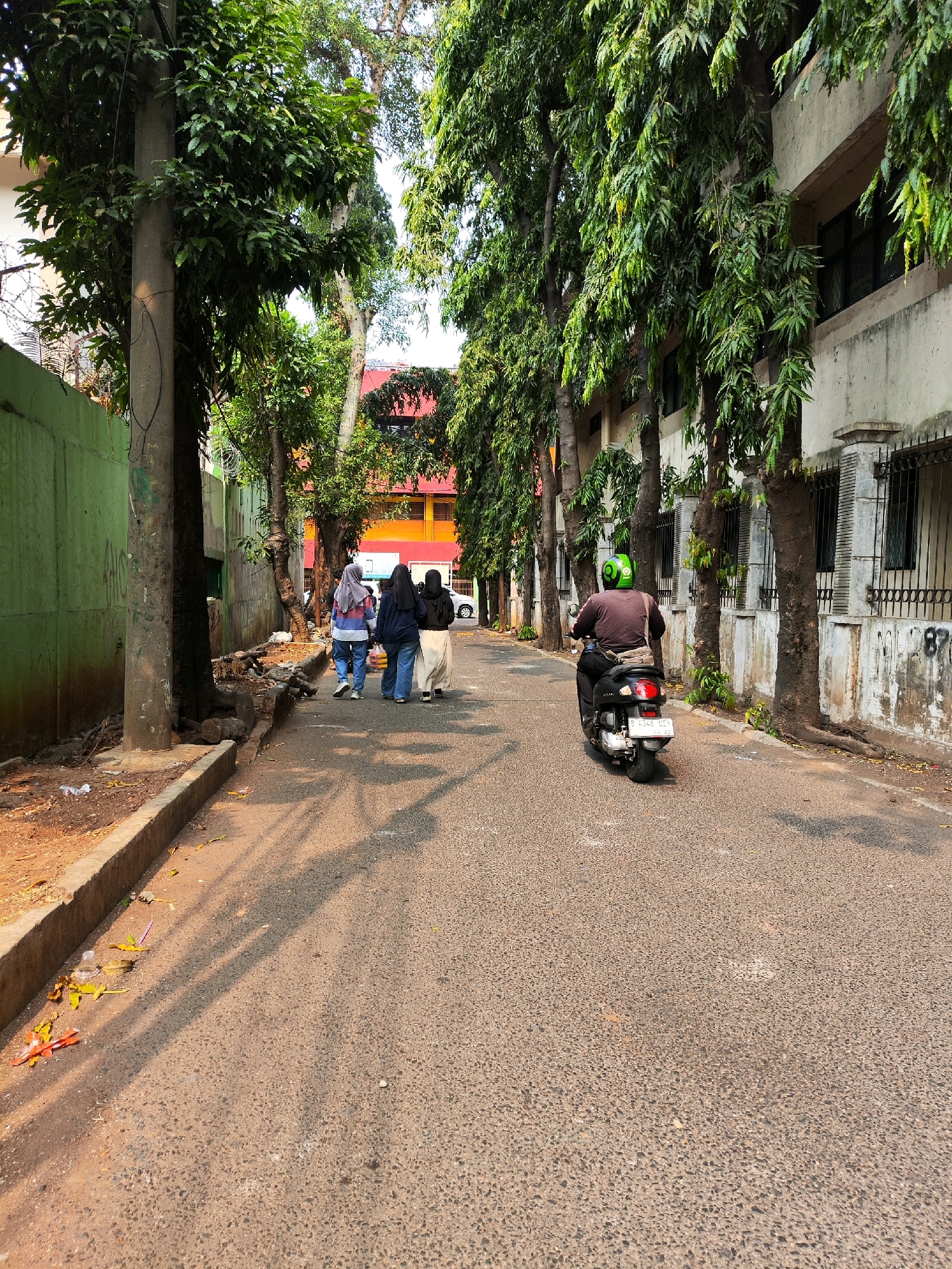 A street scene in East Jakarta, Indonesia, shows an asphalt road lined with trees. A man wearing a brown shirt and a green helmet is riding a black scooter away from the camera. Three women in hijabs are walking on the road in the same direction as the scooter. They are dressed in casual clothing. The road has a sidewalk on the left side with some debris and vegetation. Buildings with windows and balconies are visible on the right side of the road, some partially obscured by trees. The sunlight casts shadows across the road.