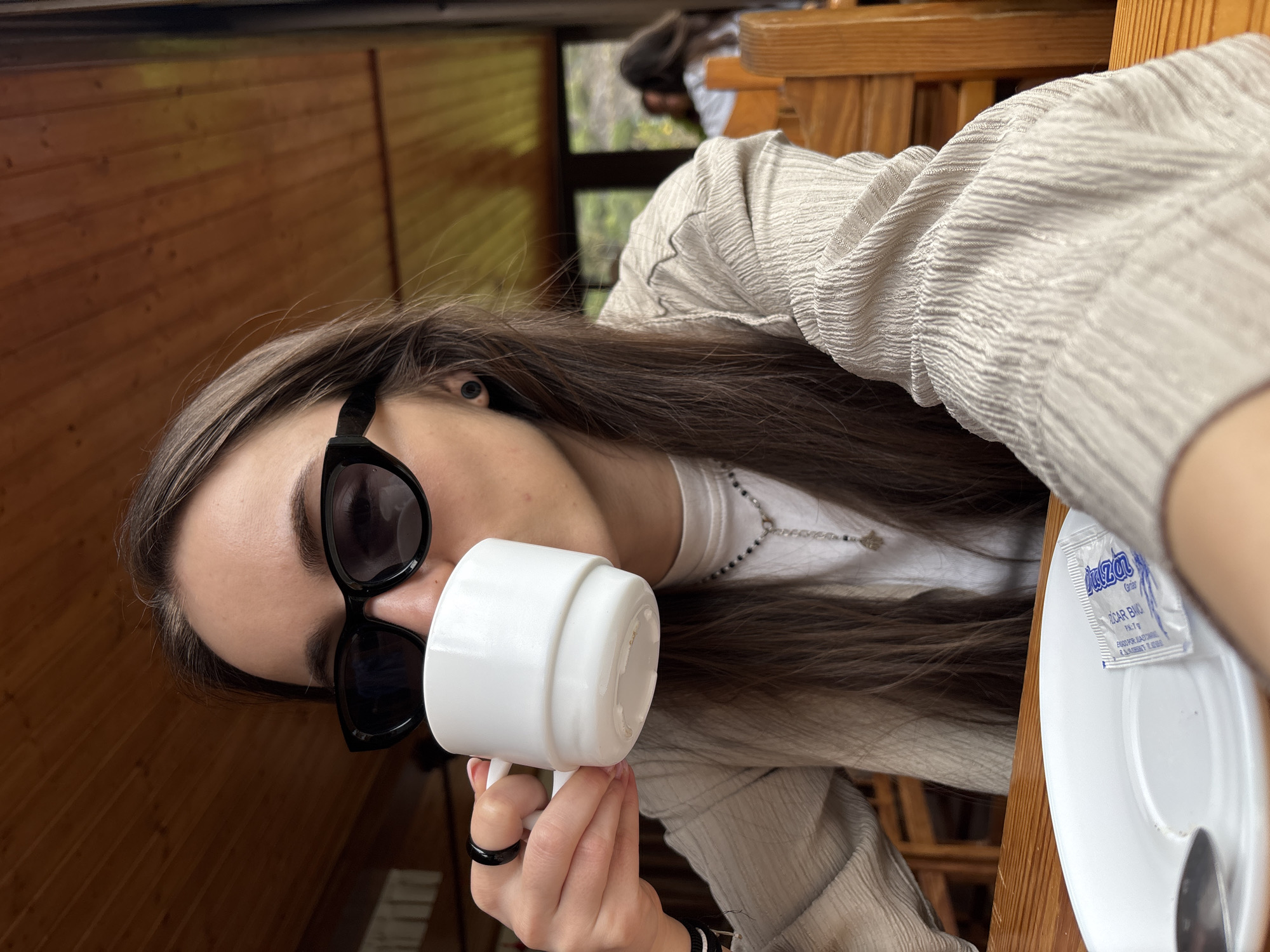 A woman with long brown hair, wearing black sunglasses, is holding a white mug to her lips. She is sitting at a wooden table, on which there is a white plate with a sugar packet and a spoon. The woman is wearing a light-colored, textured jacket and a white shirt with a necklace. The background shows a window with a glimpse of greenery and another person sitting at a table behind her. The photograph appears to be taken indoors, likely at a cafe or restaurant. The location is Buenavista del Norte, Spain.