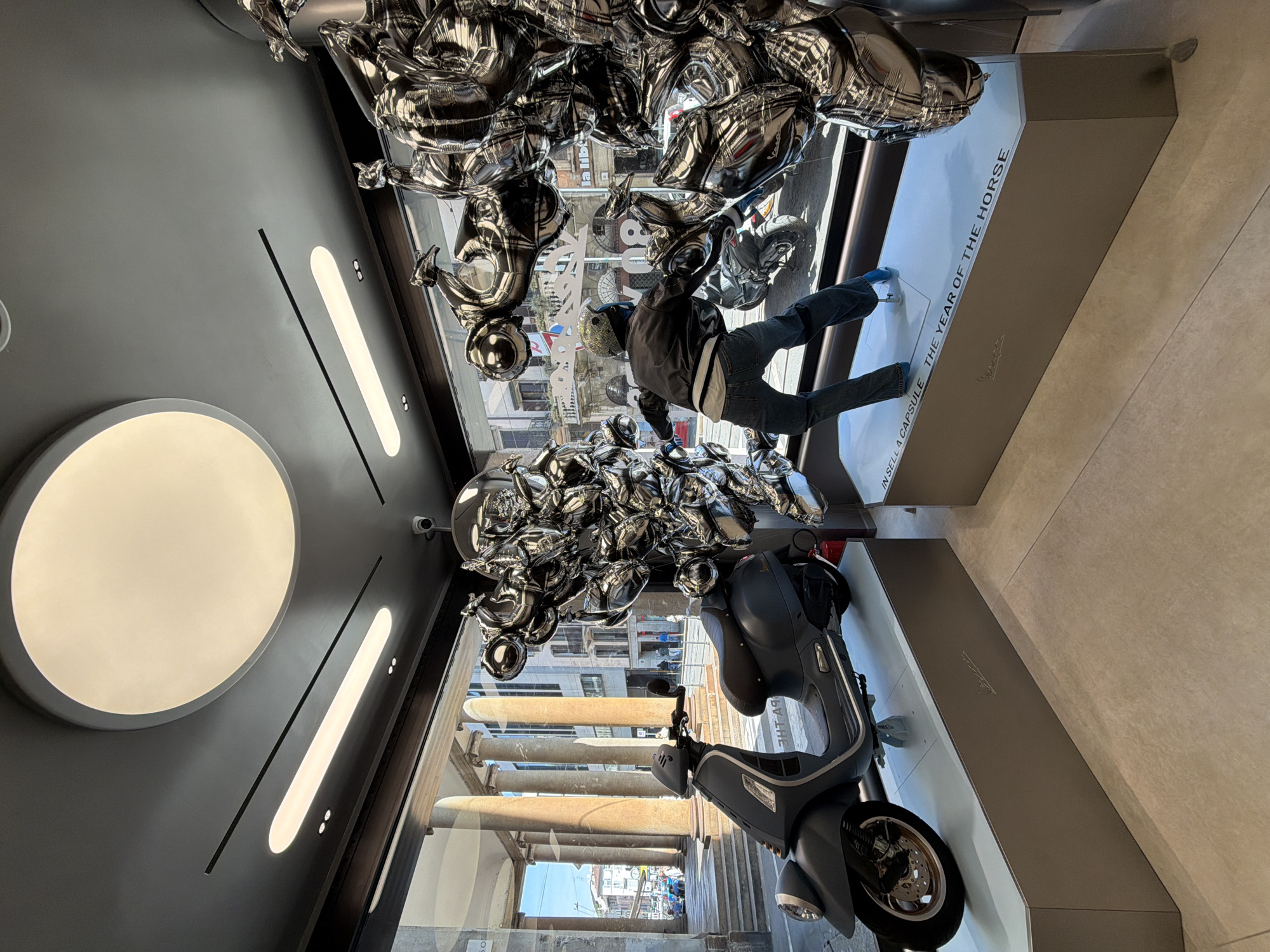 The photo captures the interior of a Vespa store in an unidentified city. A display is featured, consisting of a gray Vespa scooter mounted on a matching gray plinth which has the words "IN SELLA A CAPSULE THE YEAR OF THE HORSE". Above the scooter, silver balloon sculptures resembling Vespa scooters and riders create a whimsical, cloud-like arrangement. A mannequin dressed in riding gear is positioned with its legs projecting onto the plinth. The store's facade includes a large window displaying the streetscape outside. The outside shows the exterior of an arcade with stone columns. Inside the store, there is a gray ceiling with round lights and linear light fixtures, giving a modern and clean aesthetic.