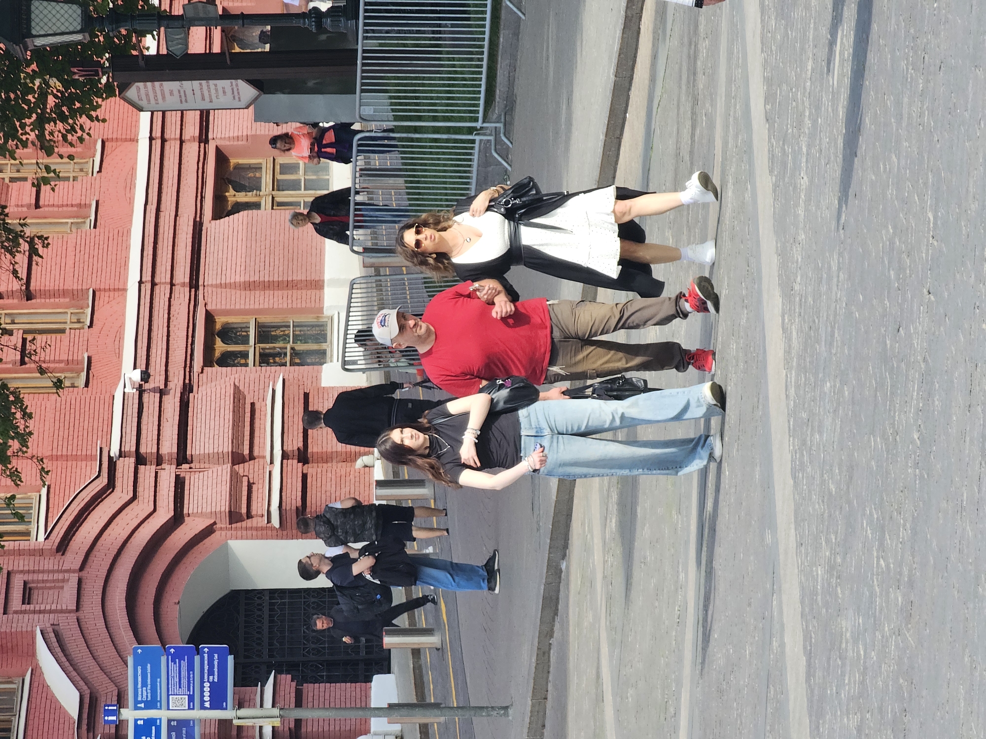 The image shows a street scene in Moscow, Russia, in front of a large red brick building with arched entrances. A signpost with blue and white directional signs is visible. Several people are walking or standing on the sidewalk. Three people are more prominently featured: a man in a red shirt, khaki pants, and a white baseball cap; a woman in a white dress with a black cardigan; and another woman in a black shirt and jeans. The background includes a metal fence and glimpses of other pedestrians. The overall scene suggests a tourist area or a public space near a historical building.