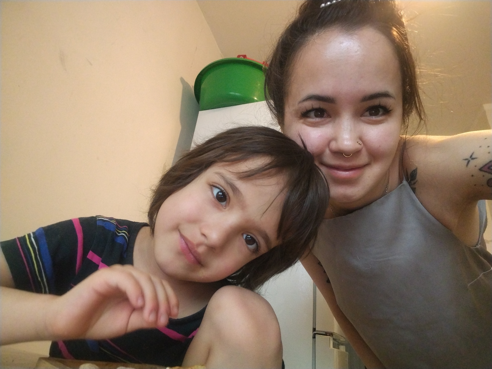 A selfie shows a woman and child indoors, likely in a kitchen, against a pale wall. The child, with brown hair, rests their head on the woman's shoulder. The woman, with dark hair pulled back, wears a nose ring and a gray top. A green container sits atop a white cabinet behind them. The location appears to be in a residential setting. There is no verifiable information that places the location specifically in the city of Noviy, Russia.