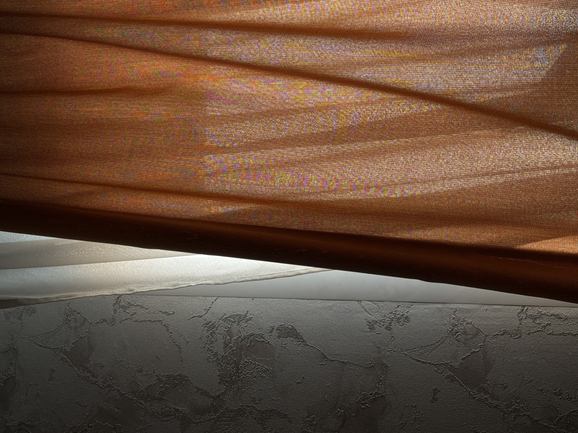 The image presents an interior scene, likely a residential room, characterized by a wall and window dressings. On the left side, a vertical section of a wall is visible, covered with textured wallpaper in a dark grey or charcoal hue. The wallpaper features a raised, irregular, and organic pattern, and this area of the wall is primarily in shadow. Adjacent to the wall, a narrow, vertical strip of translucent white or off-white sheer fabric is present, brightly illuminated from behind, indicating a light source or window. To the right, occupying approximately two-thirds of the frame, is a pleated curtain made of orange or copper-colored sheer fabric. This curtain is backlit, revealing a fine, mesh-like weave and subtle iridescent color variations (hints of pink, purple, and yellow) where light passes through. The pleats are distinct, suggesting the curtain is drawn across a window. No individuals, specific objects, or actions are observable within the frame. The precise location within Perm, Russia, cannot be determined from the visual information provided.