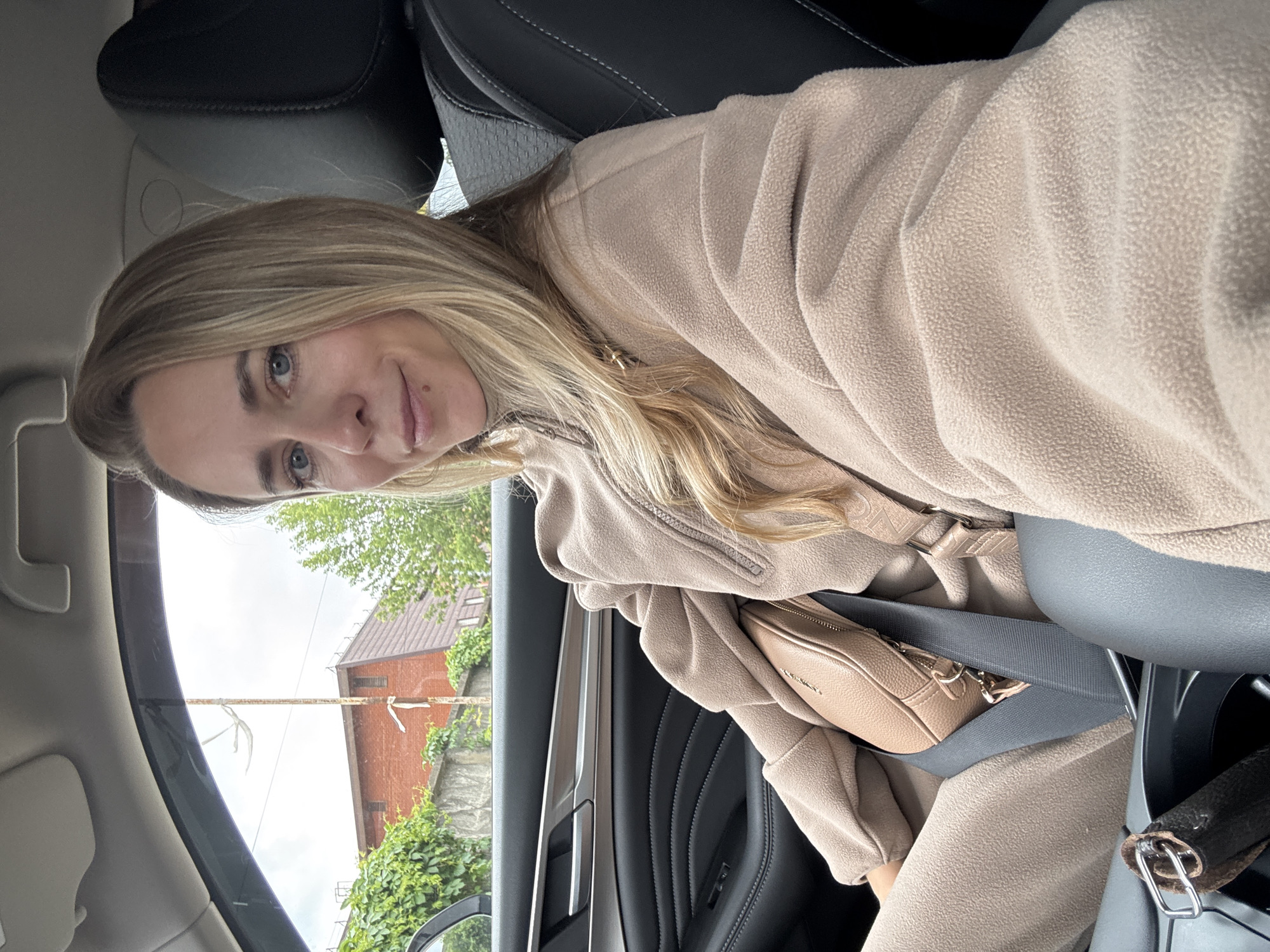 A woman is inside a car, wearing a light brown fleece jacket and a matching crossbody bag. She has long blonde hair and is wearing a seatbelt. The exterior view from the car window shows a two-story brick building with a brown roof, partially obscured by green foliage. A grey utility pole is also visible. While specific details identifying the exact location in Chaikovsky, Russia, are absent, the general architectural style suggests a residential or commercial area within the city.
