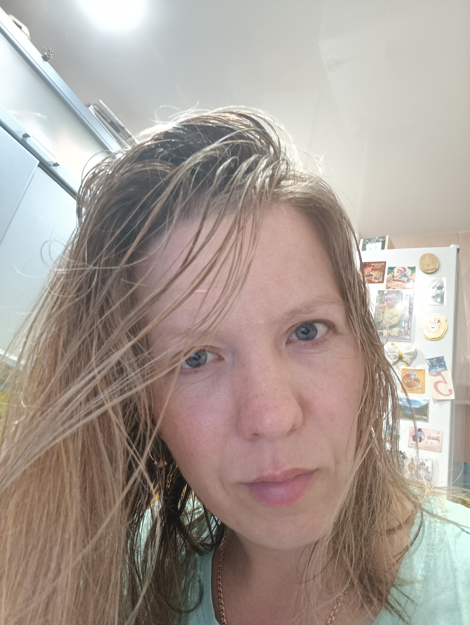 The image is a close-up selfie of a woman with wet, blonde hair. Her eyes are blue and she has fair skin with freckles. She is wearing a light green shirt and a gold necklace. The background suggests a kitchen setting, with a white refrigerator covered in magnets and pictures visible behind her. The location is likely Chaikovsky, Russia.