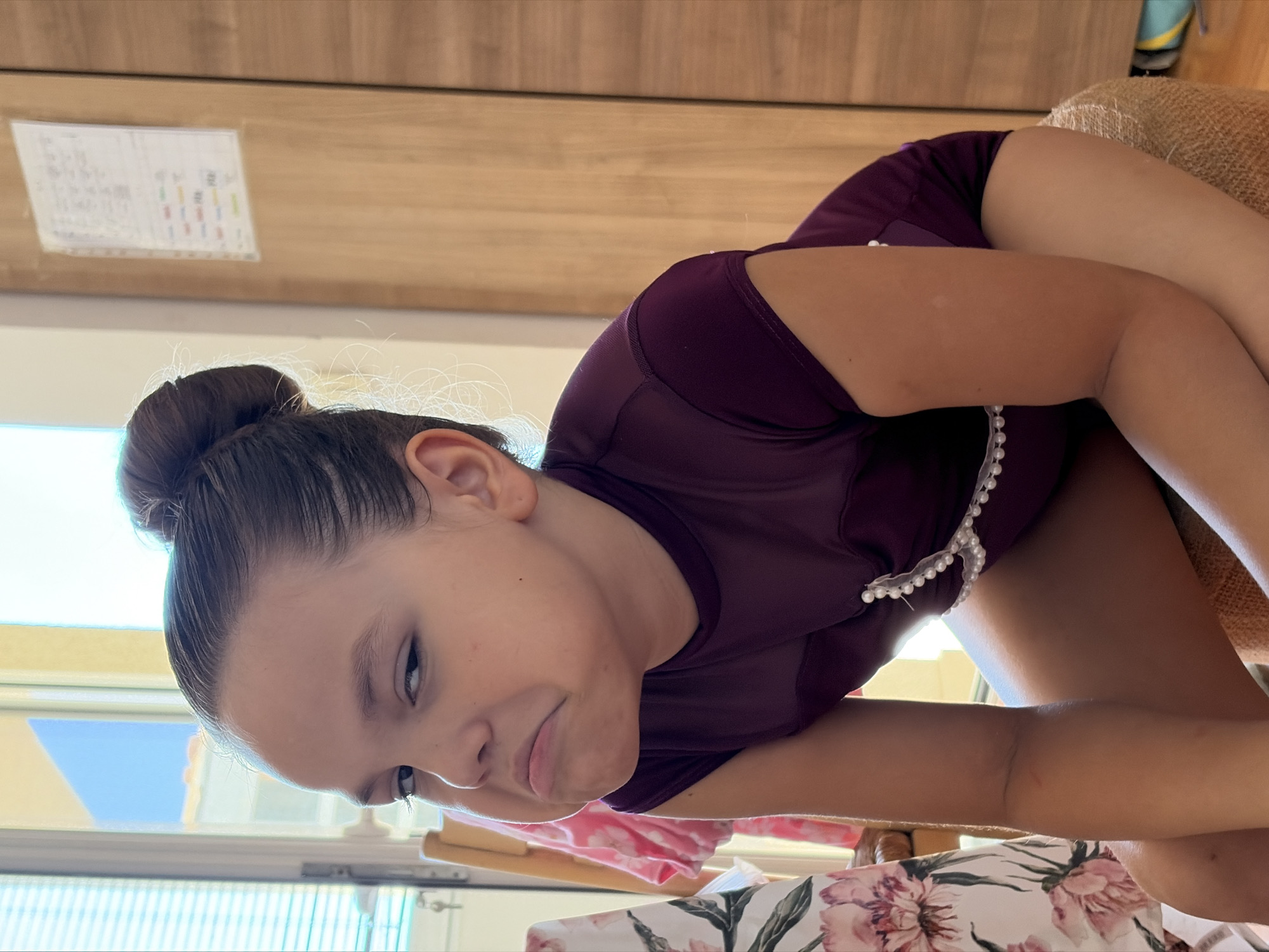 A young girl with her hair pulled into a bun is shown in a close-up, angled shot. She is wearing a dark purple leotard with a decorative pearl trim along the neckline. Her expression is somewhat downcast, with her head tilted. The background consists of a wooden cabinet and a window with a view of the outdoors, suggesting an indoor setting. A printed fabric with floral patterns is visible in the foreground. There is a piece of paper with some writing and colorful squares attached to the wooden cabinet in the upper left portion of the image. The lighting suggests it is daytime. The girl appears to be in a relaxed or resting pose.